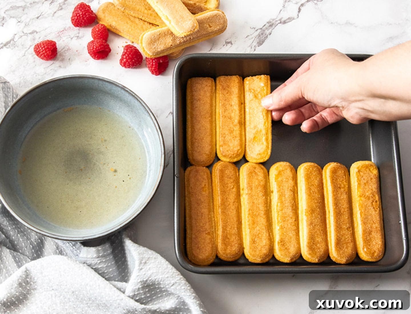 A hand gently placing liqueur-dipped ladyfingers into a rectangular metal baking pan, forming the foundational layer of the tiramisu.