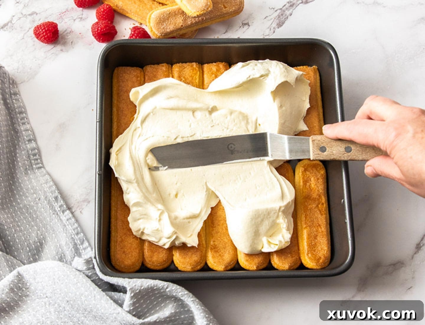 An offset spatula expertly spreading the smooth, creamy raspberry tiramisu filling over the first layer of ladyfingers in a baking dish.
