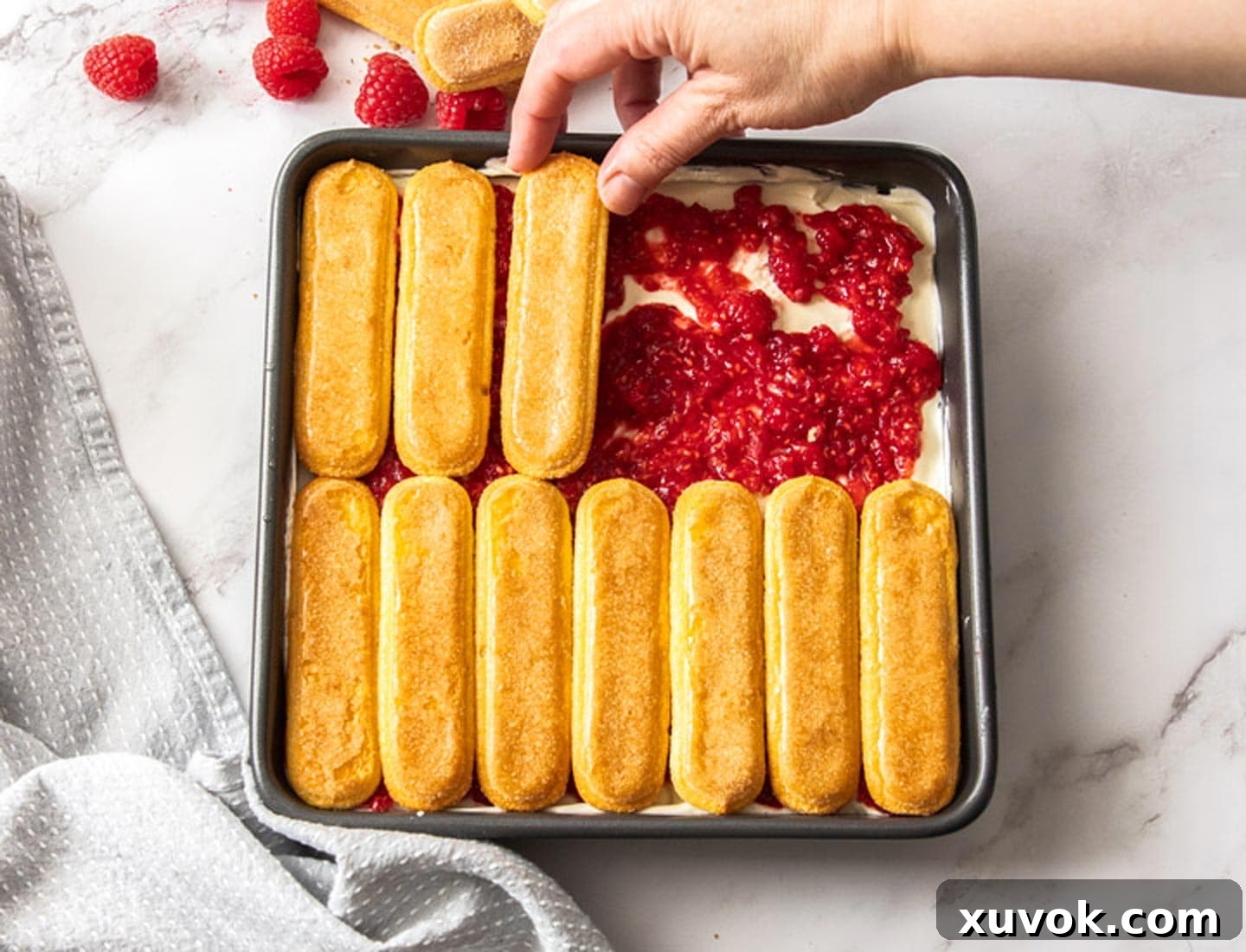 A hand carefully adds a second layer of liqueur-dipped ladyfingers on top of the mashed raspberries, continuing the tiramisu assembly.