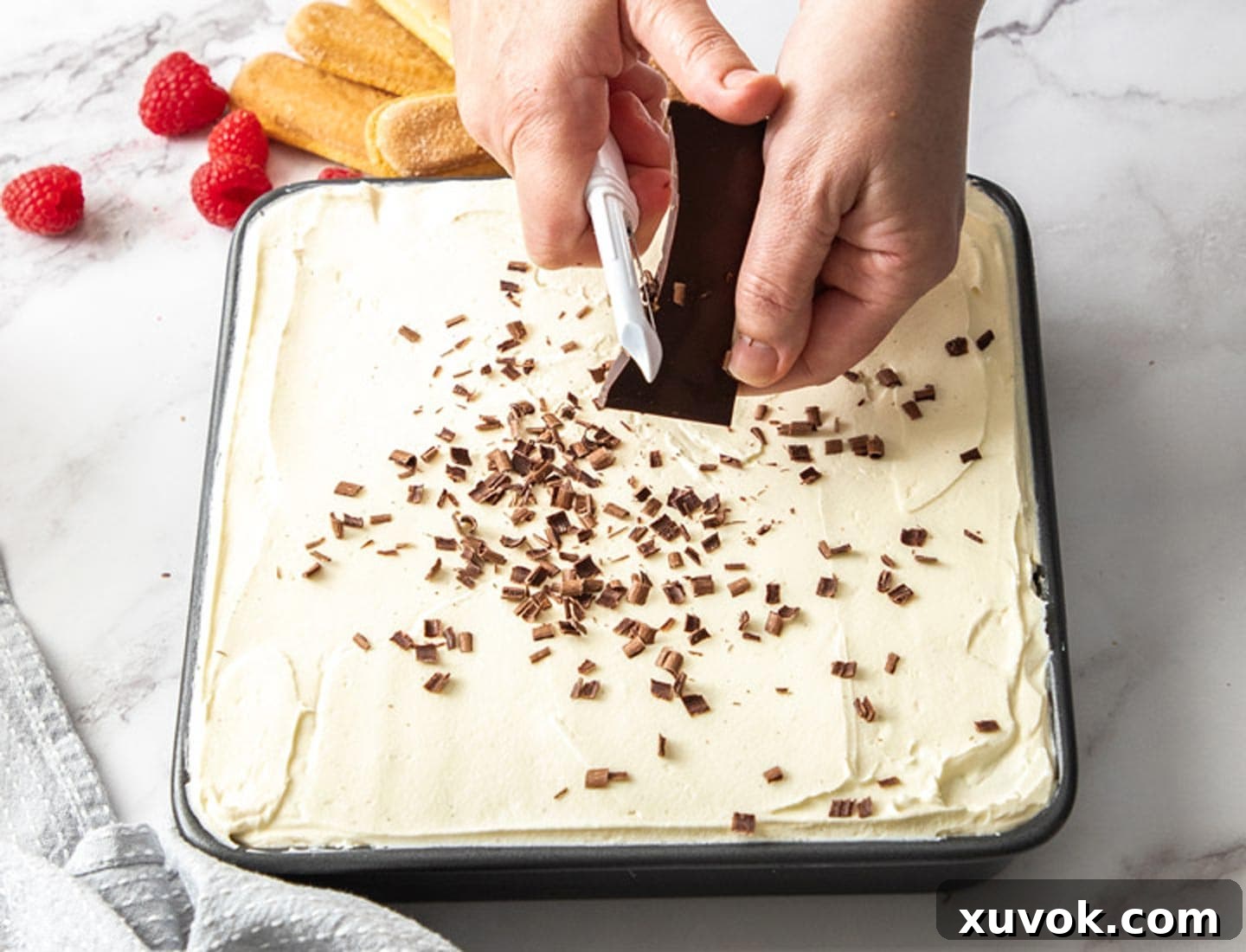 Two hands meticulously using a vegetable peeler to shave dark chocolate directly onto the top of the creamy raspberry tiramisu, adding the final garnish.