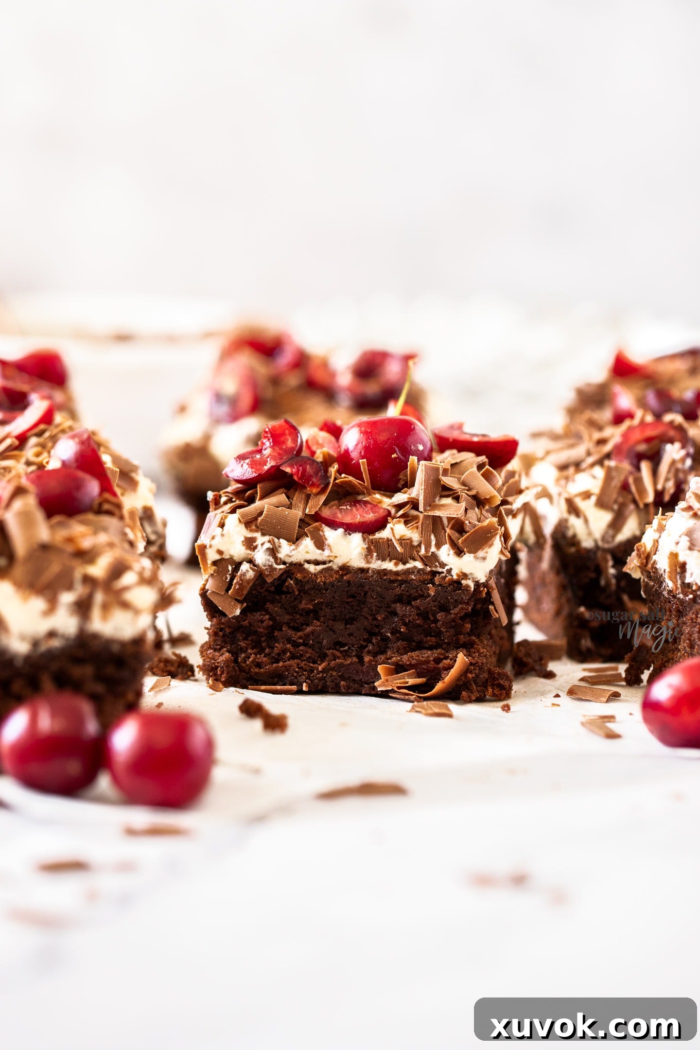 A rich, fudgy Black Forest Brownie topped with a generous swirl of whipped cream, fresh red cherries, and delicate chocolate shavings, resting on a clean white surface. The close-up shot highlights the luxurious texture and vibrant colors.