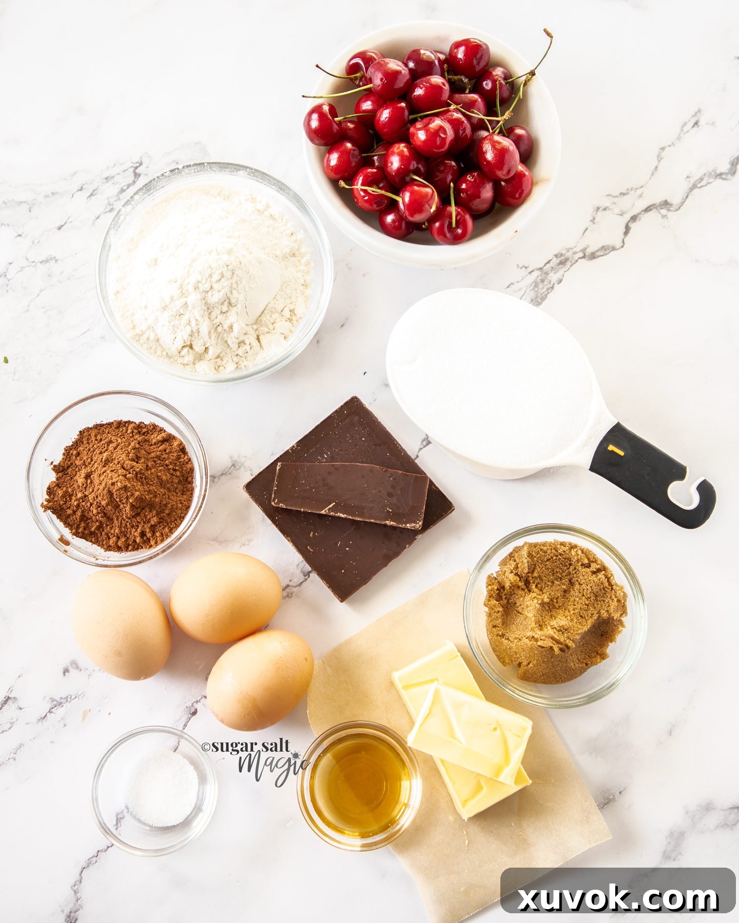 A collection of fresh and pantry ingredients laid out on a white marble surface, ready for making Black Forest Brownies. Includes fresh cherries, dark chocolate, butter, eggs, flour, cocoa powder, and sugars.