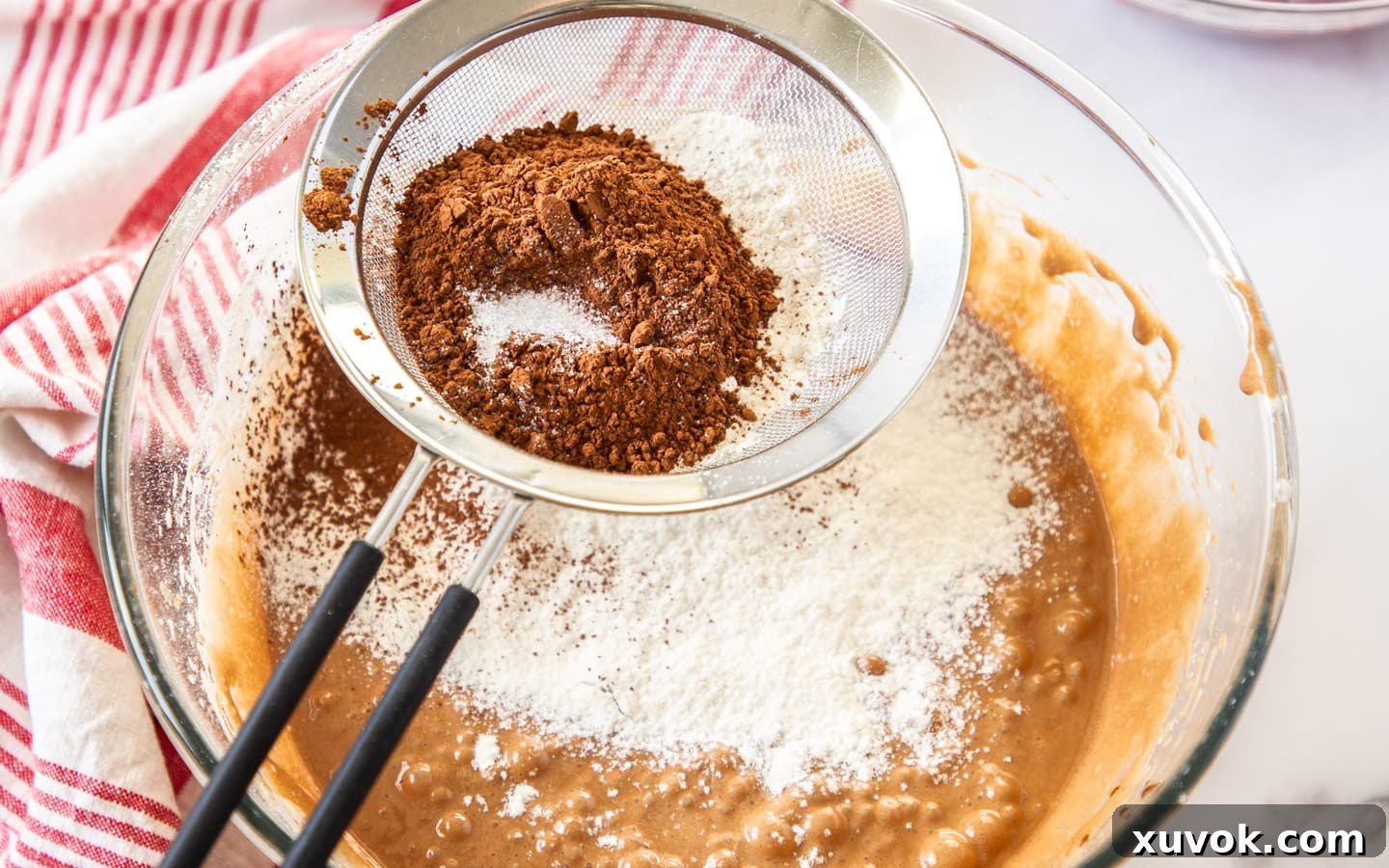 A fine mesh sieve sifting plain flour, cocoa powder, and salt over the rich chocolate brownie batter in a mixing bowl.