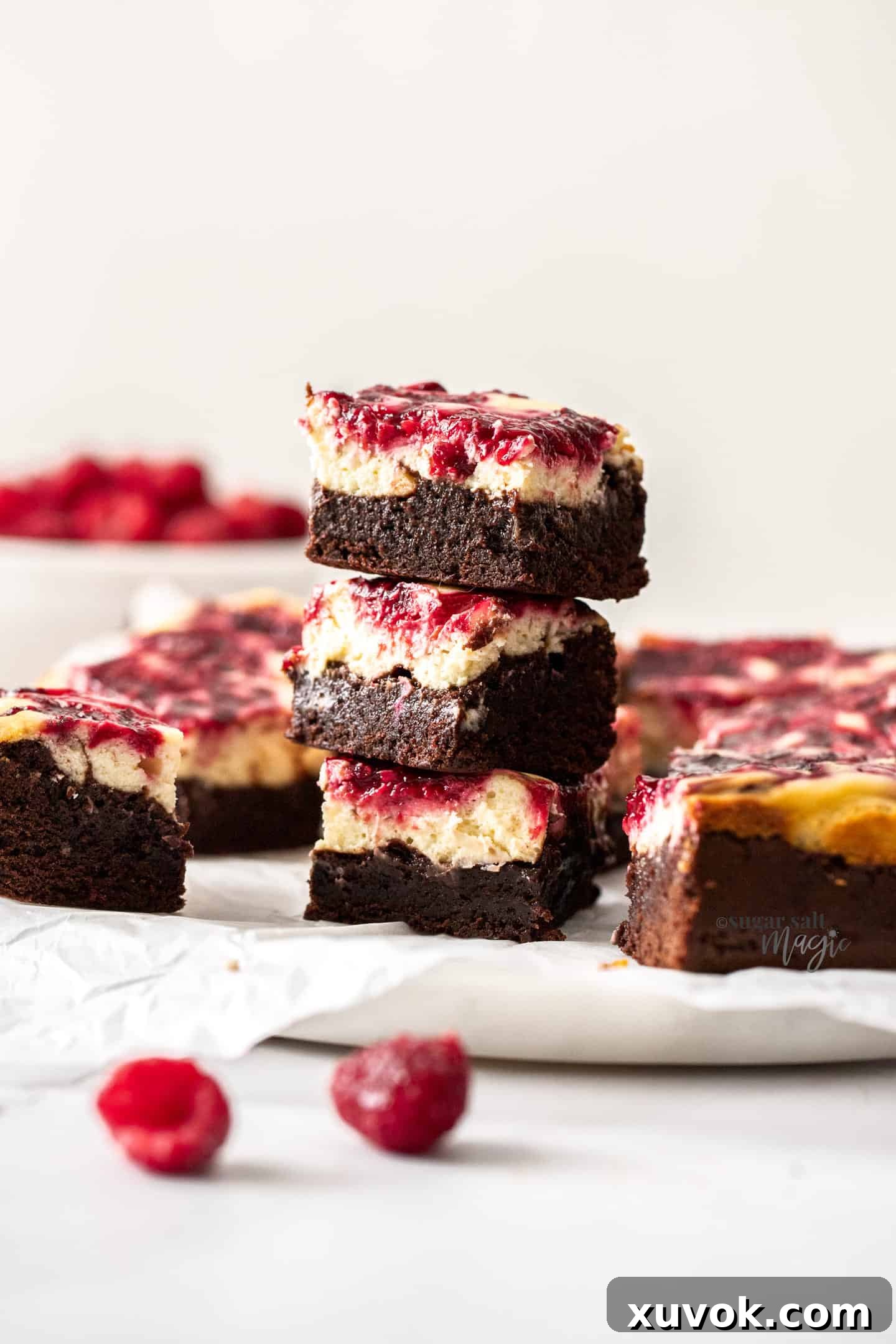 A beautifully presented stack of three raspberry cheesecake brownies, garnished with fresh raspberries, on an elegant marble cake plate.