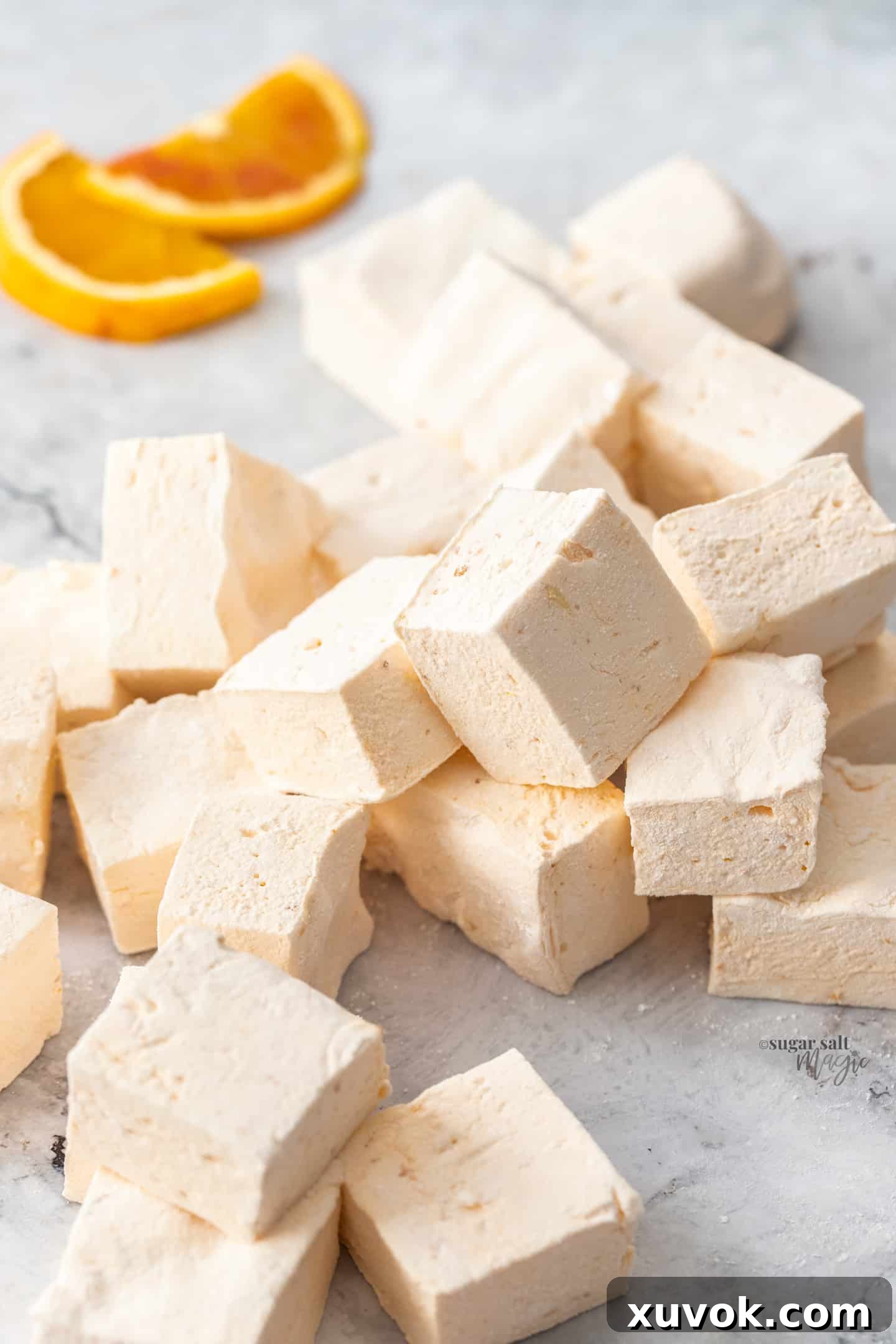 A generous pile of homemade orange marshmallows, dusted with powdered sugar, resting on a sophisticated grey marble surface. Each marshmallow is soft, squishy, and light as air, brimming with a festive orange flavor.