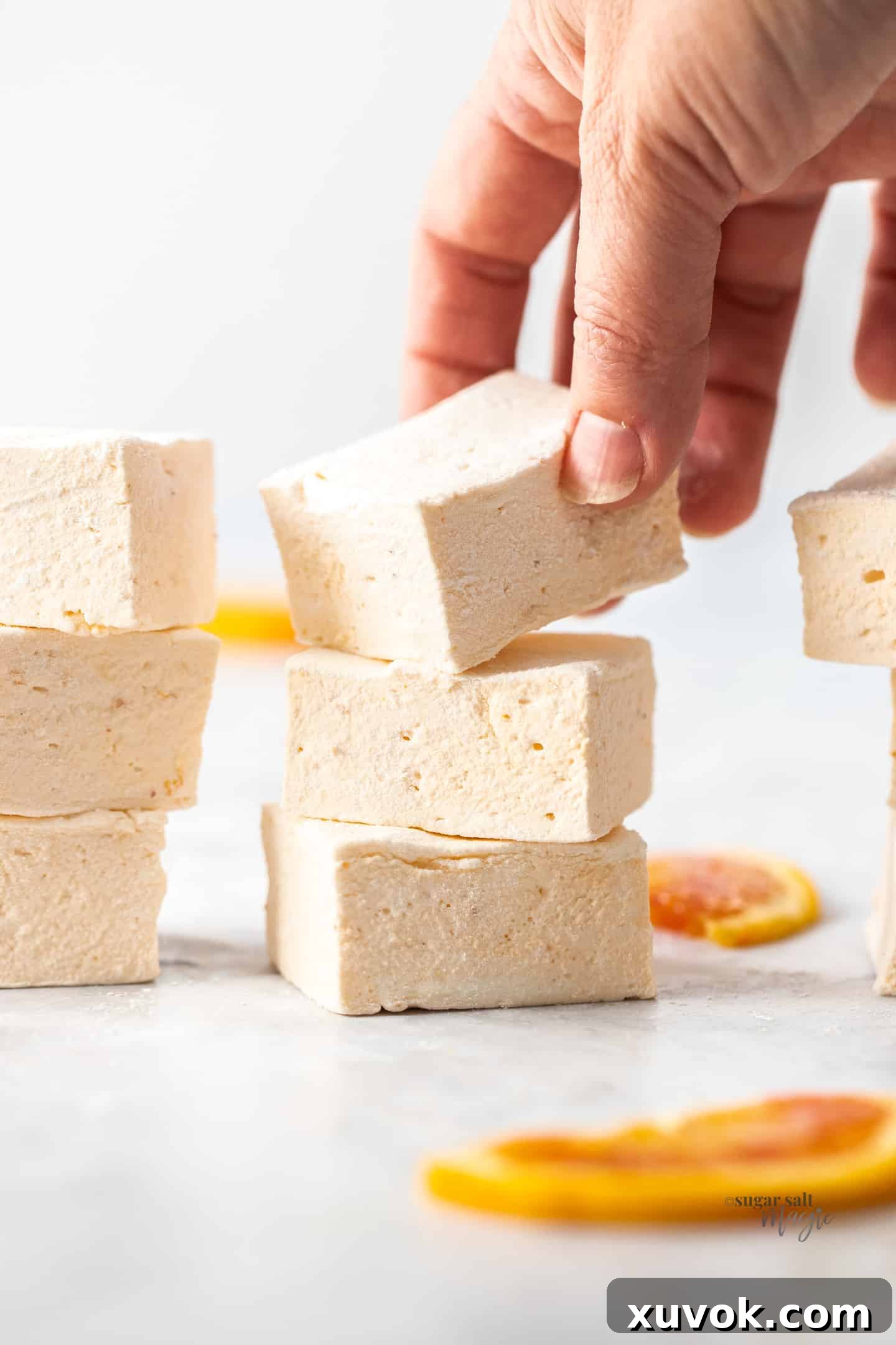 A hand reaches out to pick a marshmallow from a stack of three vibrant orange marshmallows, all dusted with powdered sugar, set against a grey marble background. The marshmallows appear soft and inviting.