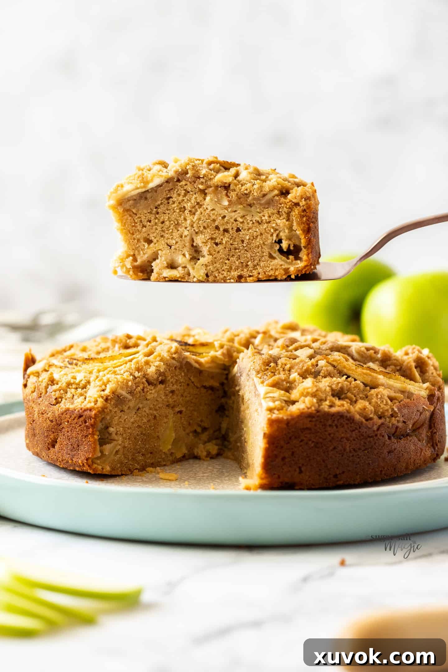 A delectable slice of Cinnamon Apple Crumb Cake being lifted, showcasing the moist cake, tender apples, and crunchy streusel topping. The background features the rest of the beautifully baked cake.