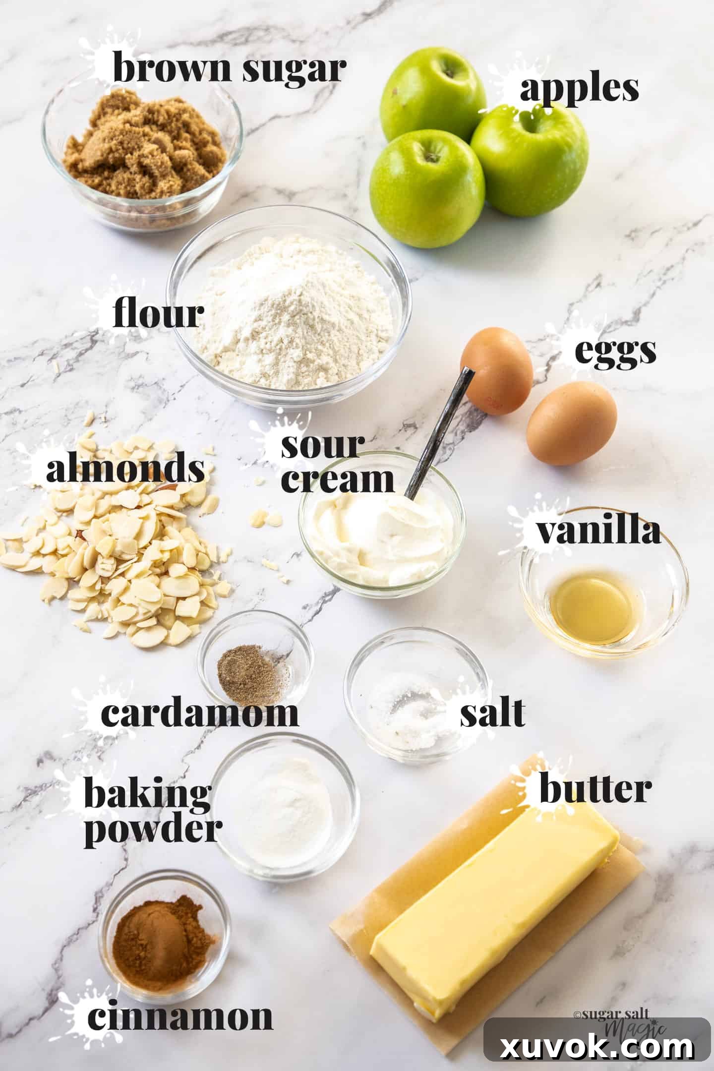 A curated display of fresh ingredients for baking apple crumb cake, including apples, flour, sugar, butter, and spices, arranged artfully on a marble countertop.