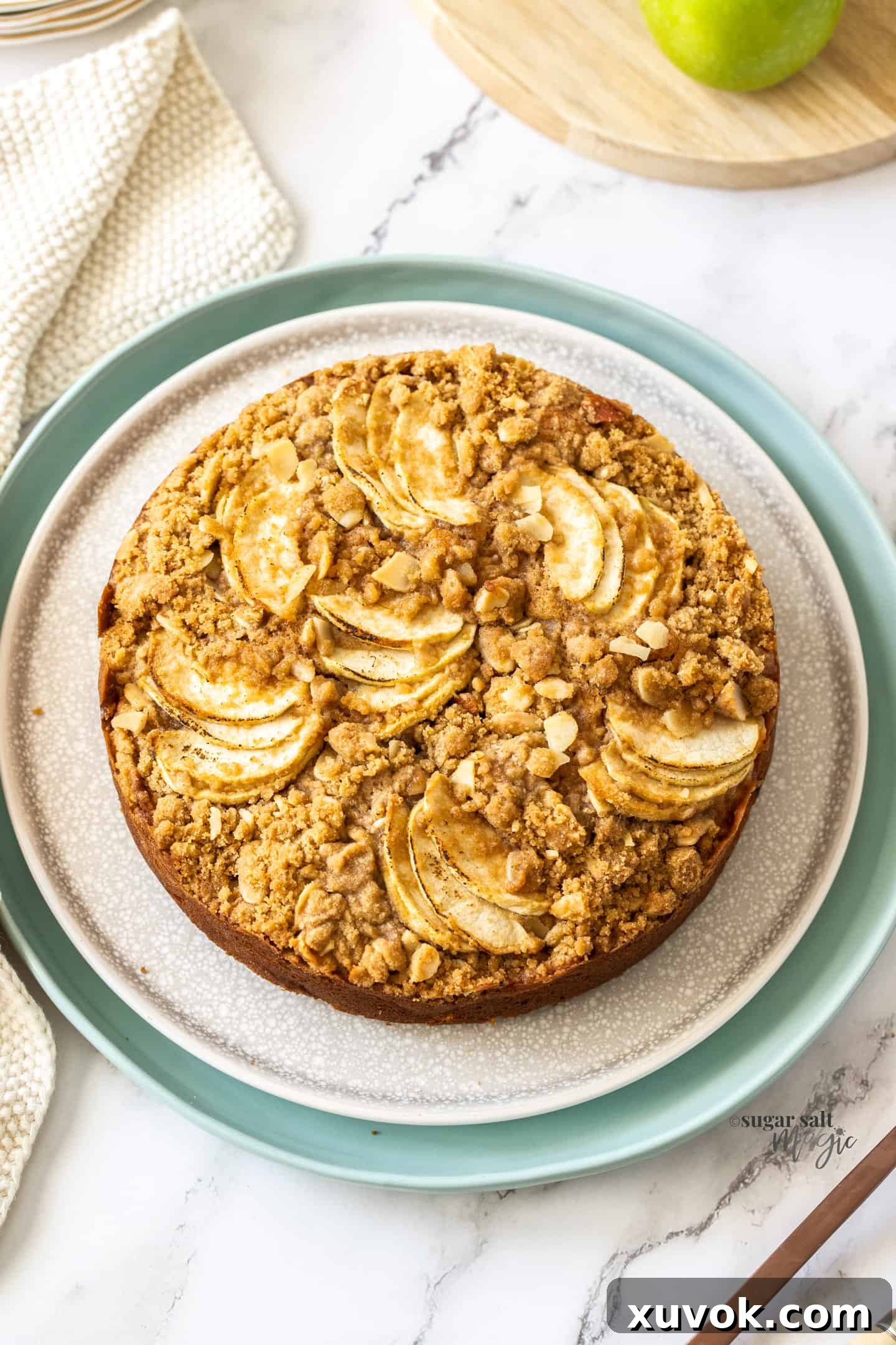 A top-down view of a freshly baked Cinnamon Apple Crumb Cake, golden brown with a generous crumb topping, presented on a rustic cake stand.