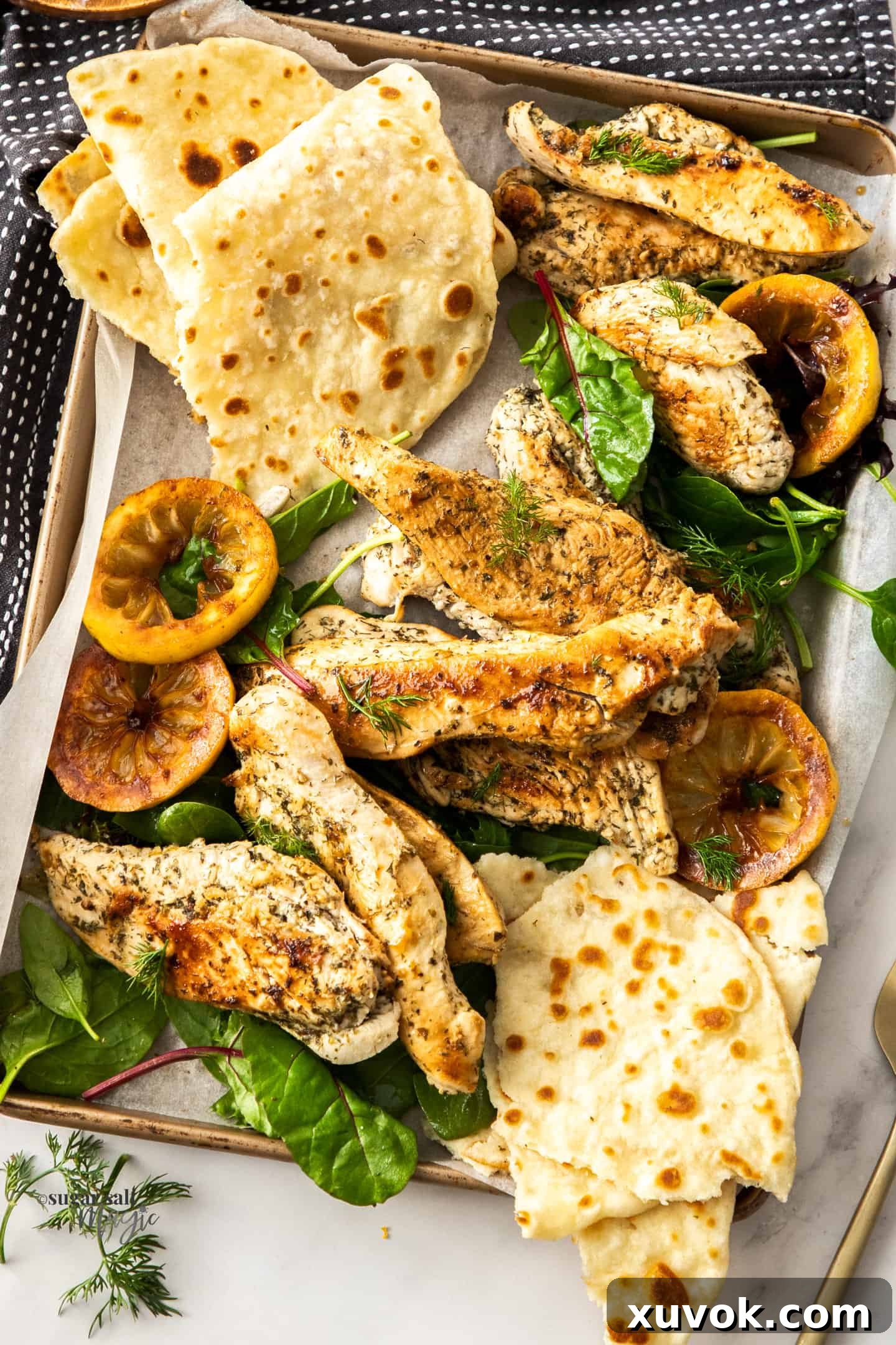 Top down view of a tray filled with cooked Greek seasoned chicken pieces and a fresh, colorful salad, garnished with vibrant lemon slices and fresh herbs. The chicken is golden brown and looks juicy.