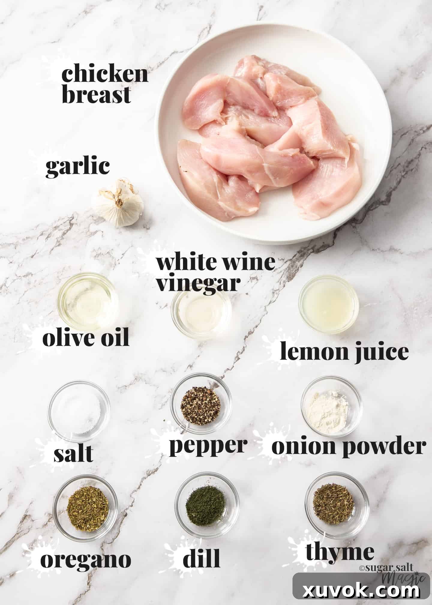 Ingredients laid out on a white marble surface for making Greek chicken, including fresh lemon, a bottle of olive oil, a bowl of minced garlic, and small dishes of various dried herbs like oregano, dill, and thyme.