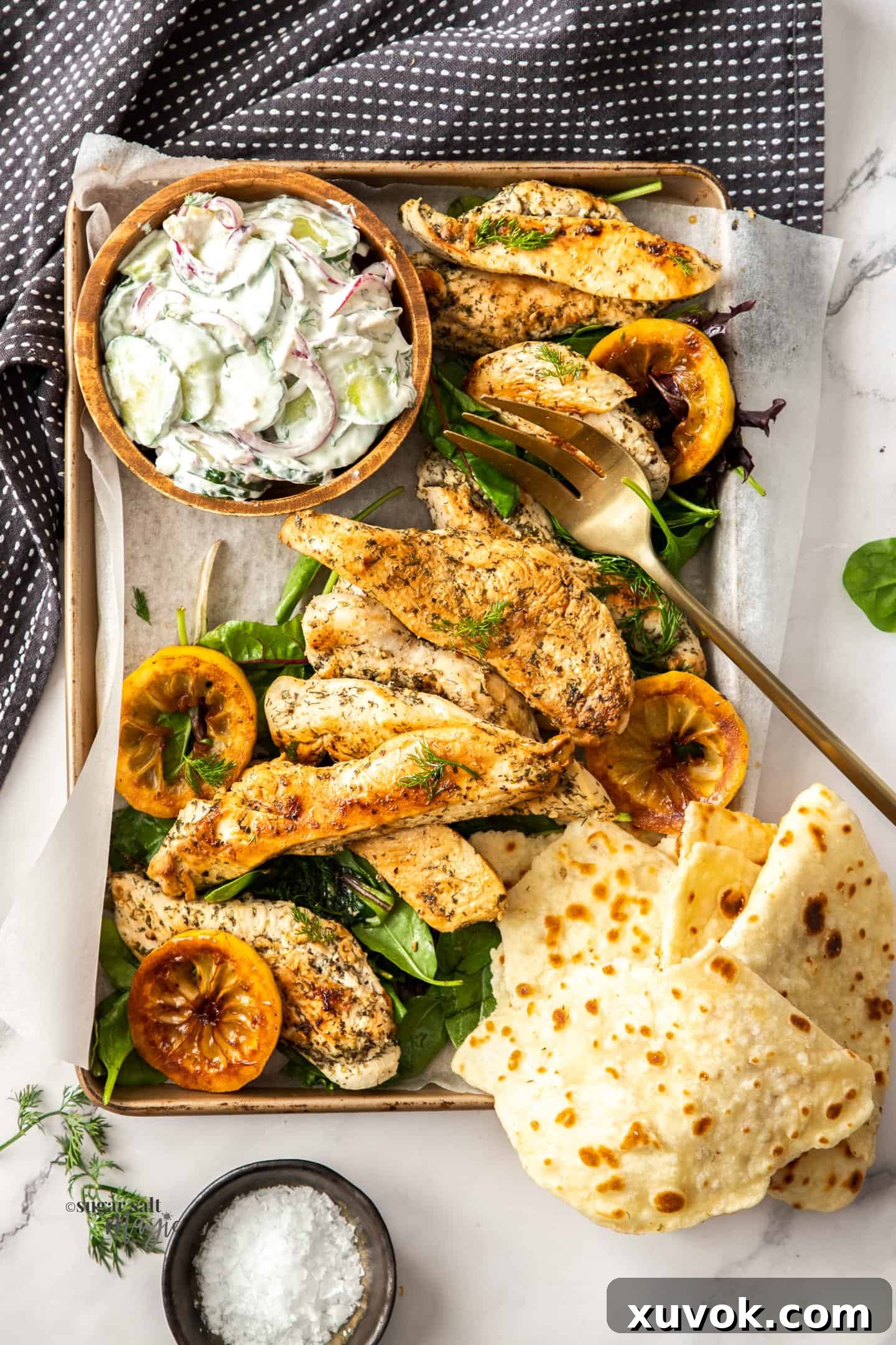 Top down view of a tray filled with perfectly cooked Greek seasoned chicken pieces and a vibrant fresh salad, garnished with lemon slices and herbs, all beautifully arranged and ready for serving.