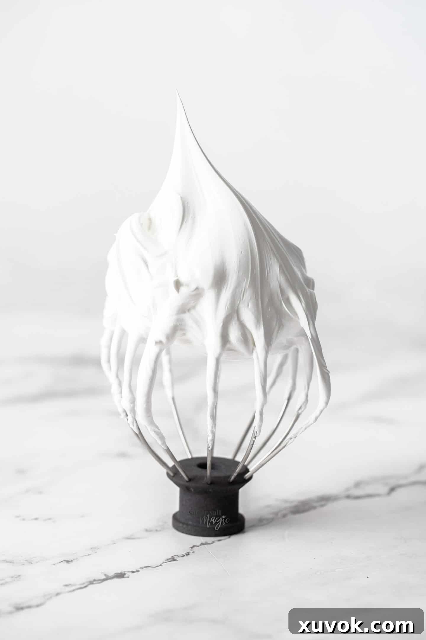 A whisk standing upright with a peak of meringue on top.