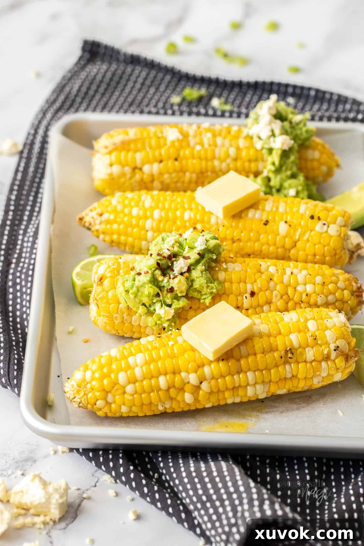 Four cobs of perfectly cooked air fryer corn on a silver baking tray, lightly topped with butter and avocado slices.