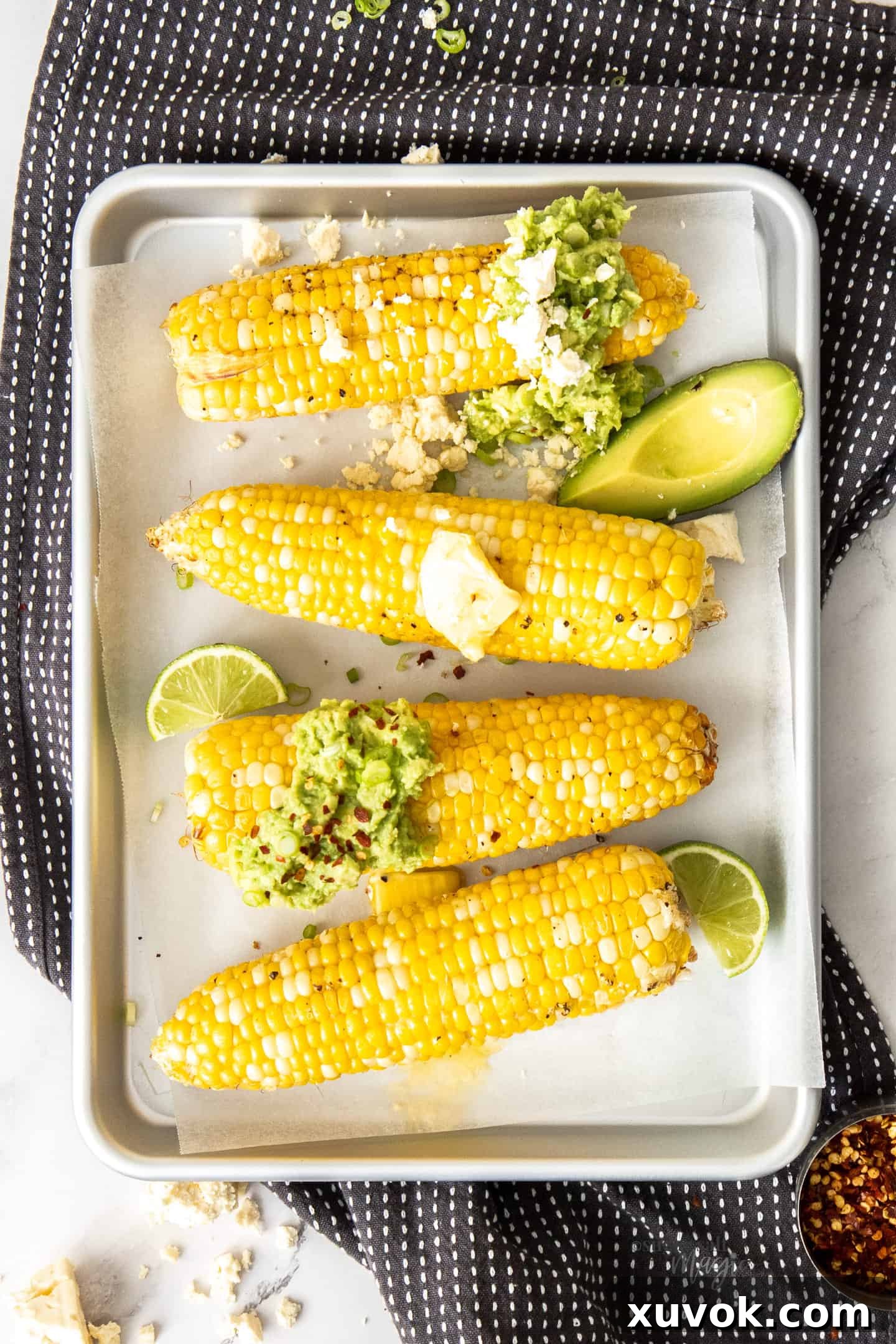 Four cobs of air-fried corn on a silver baking tray, artfully garnished with pats of butter and fresh avocado slices, ready to be enjoyed.