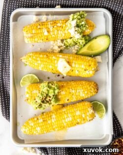 Four cobs of corn on a silver baking tray topped with butter and avocado.