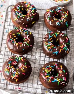 Six chocolate sprinkle donuts arranged on a wire rack with a bowl of colorful sprinkles in the background, a perfect sweet treat.
