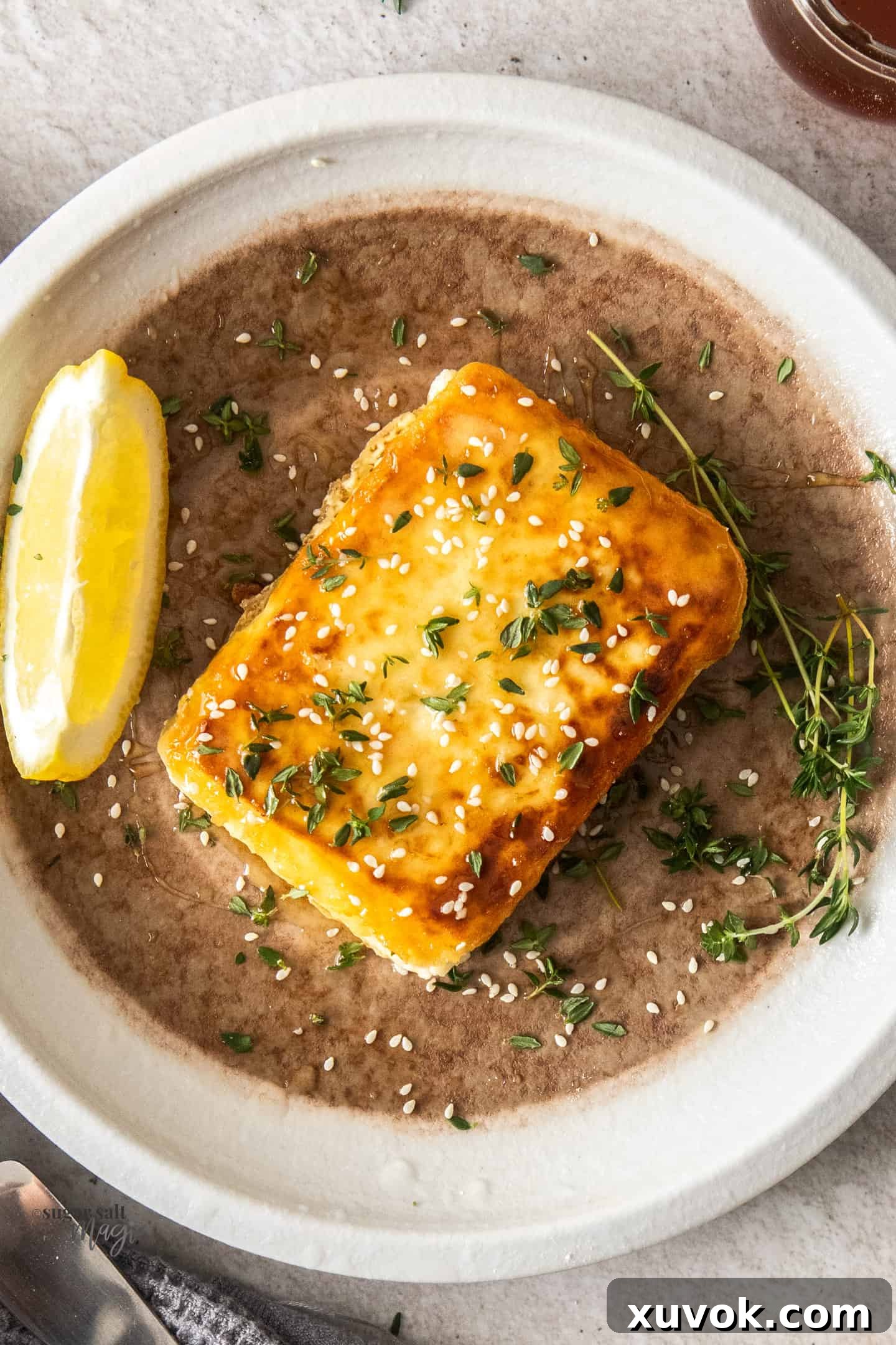 Top down view of a slice of fried feta on a plate with a lemon wedge on the side, drizzled with honey and sprinkled with thyme and sesame seeds.