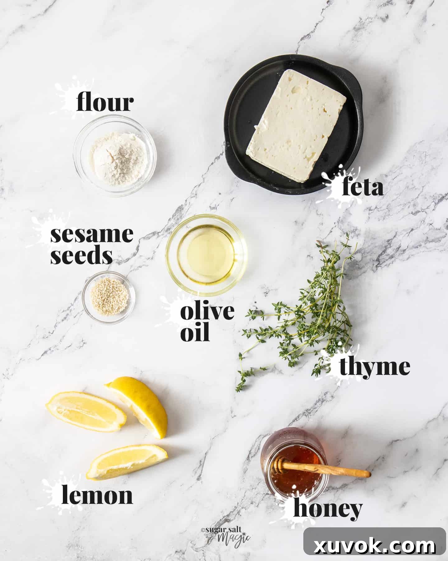 Ingredients for feta saganaki artfully arranged on a marble benchtop, including a block of feta, olive oil, flour, lemon, honey, thyme, and sesame seeds.