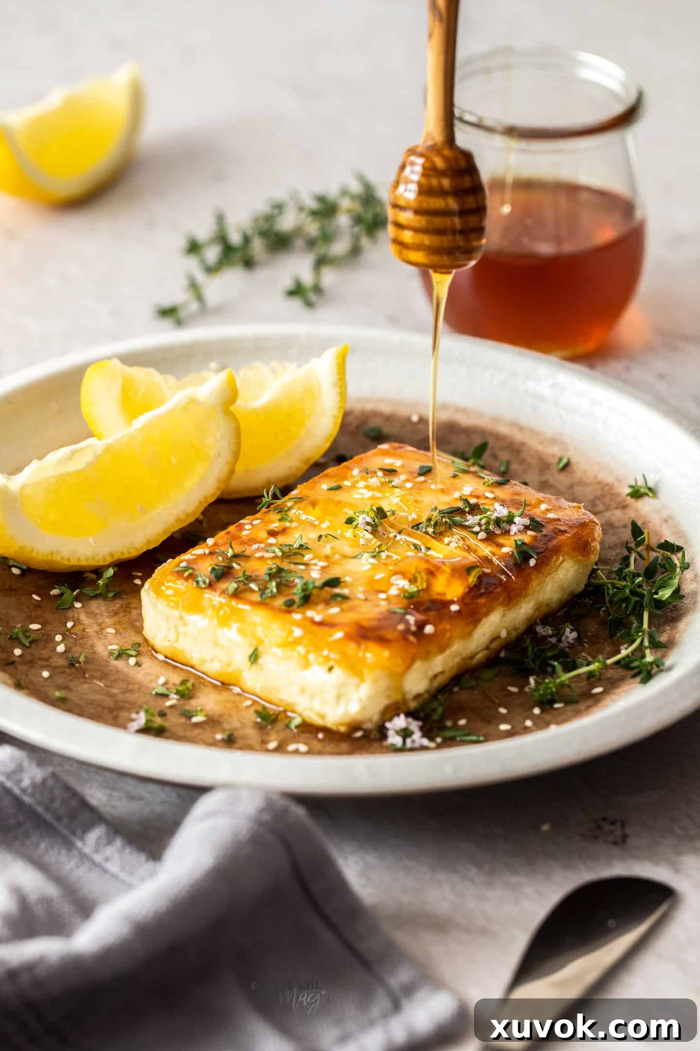 Honey being drizzled onto a perfectly pan-fried square of feta cheese, with fresh thyme leaves and sesame seeds scattered on top.