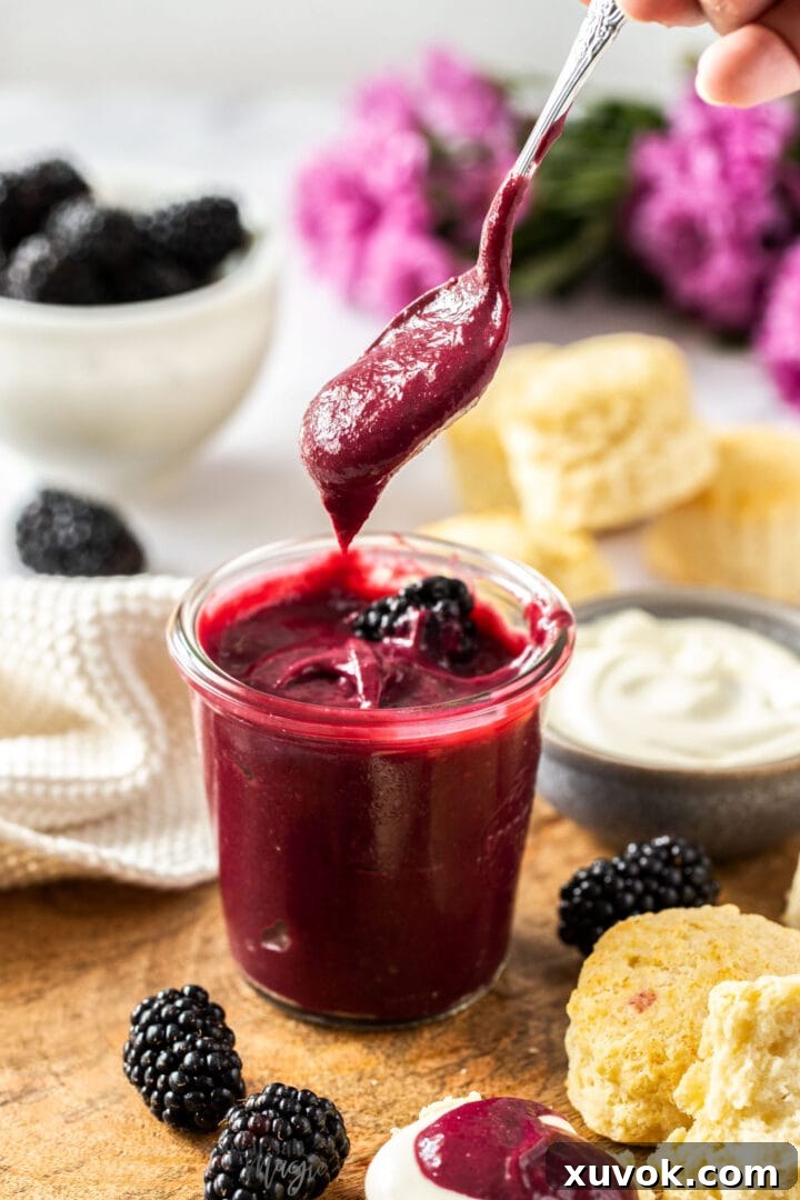 A spoon delicately positioned above a glass jar filled with luscious homemade blackberry curd, showcasing its rich, glossy texture and inviting appeal, ready to be enjoyed as a spread or filling.