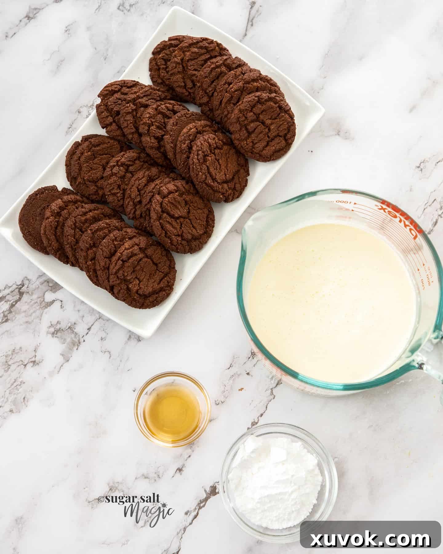 Four core ingredients for chocolate ripple cake – a pack of chocolate biscuits, a carton of cream, a bowl of icing sugar, and a bottle of vanilla extract – neatly arranged on a marble surface.