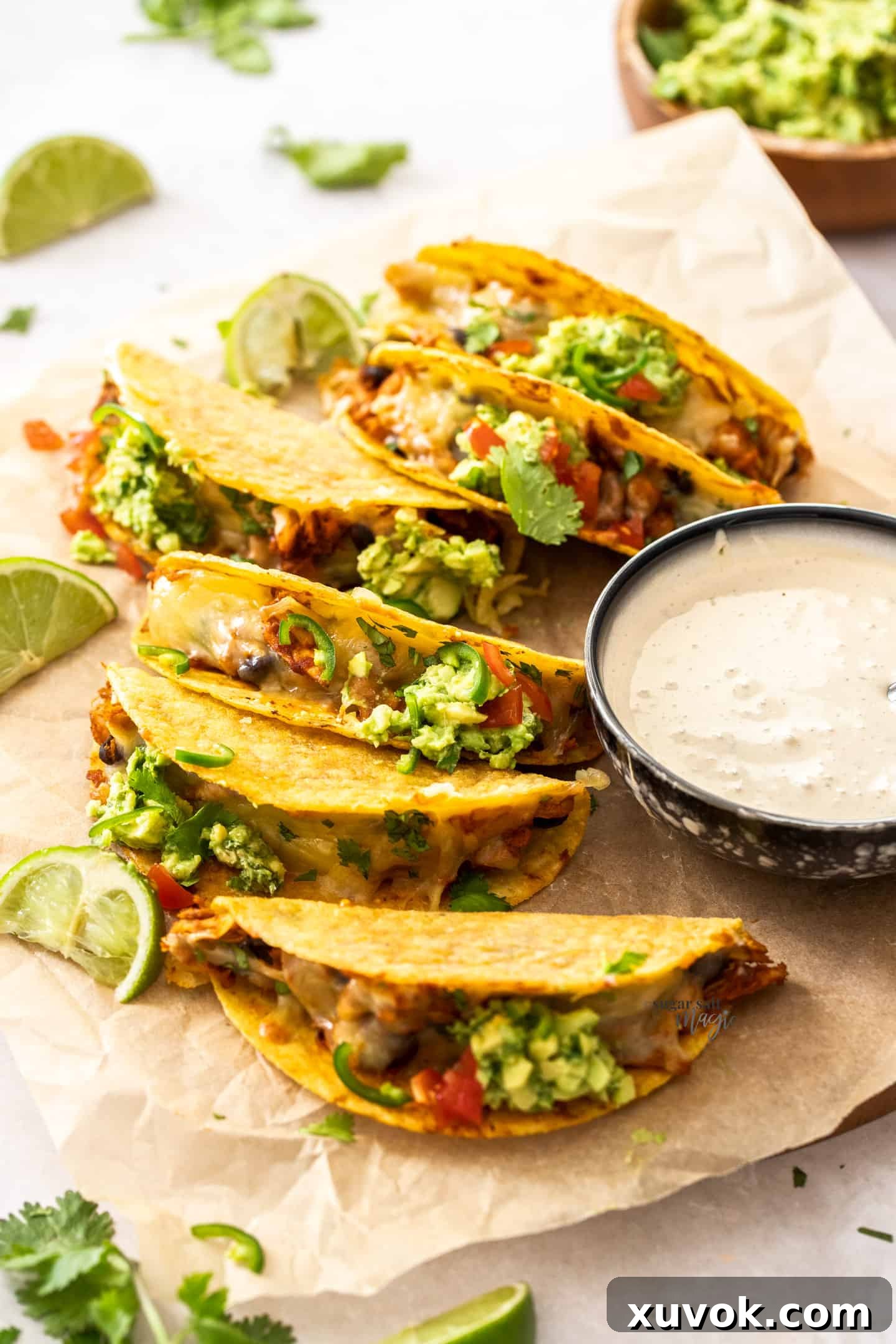 A batch of golden-brown oven-baked chicken tacos lined up on a sheet of baking paper, filled with chicken and melted cheese, ready to be topped.