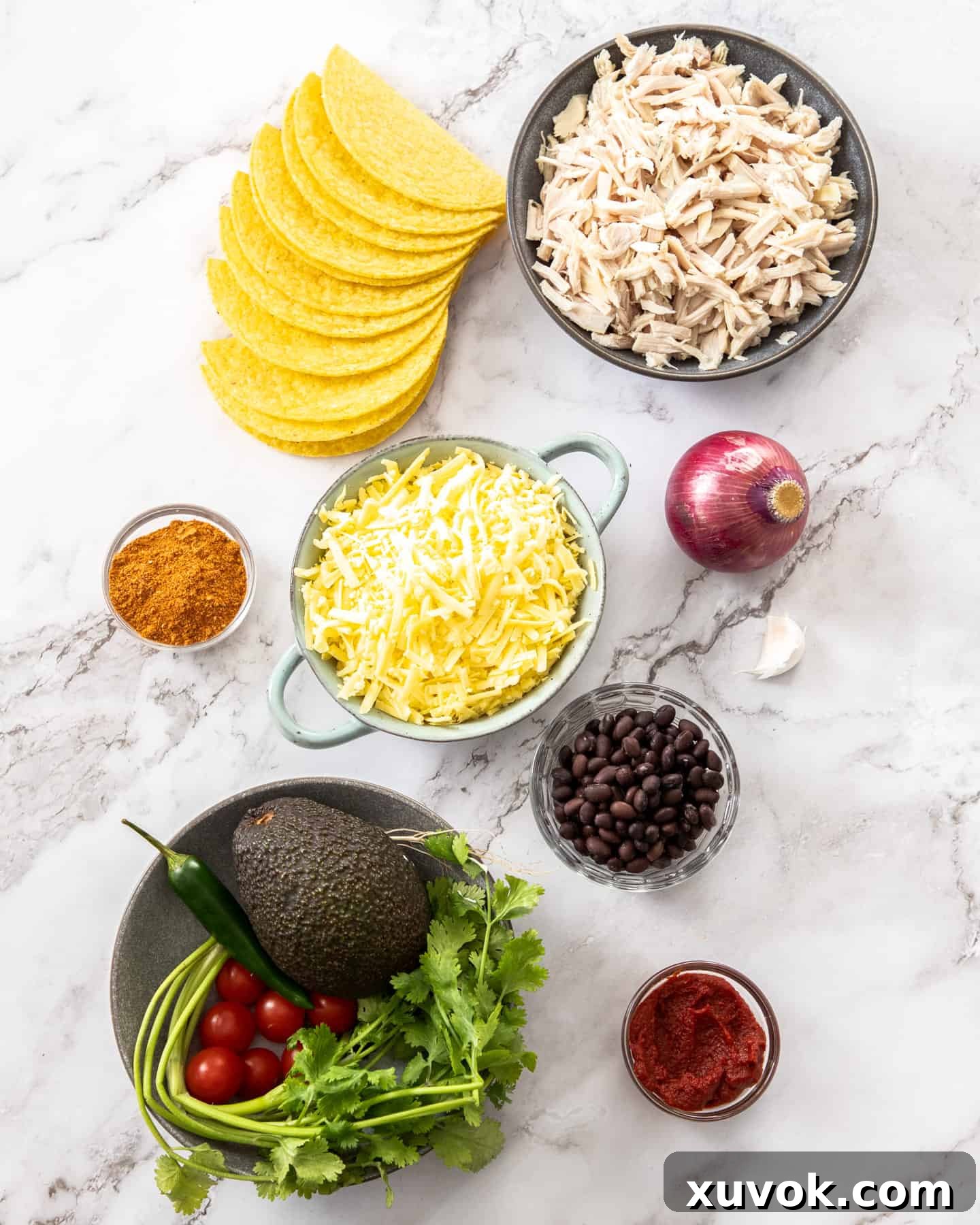 Various ingredients for baked chicken tacos are neatly arranged on a white marble surface, including shredded chicken, taco shells, cheese, and fresh produce.