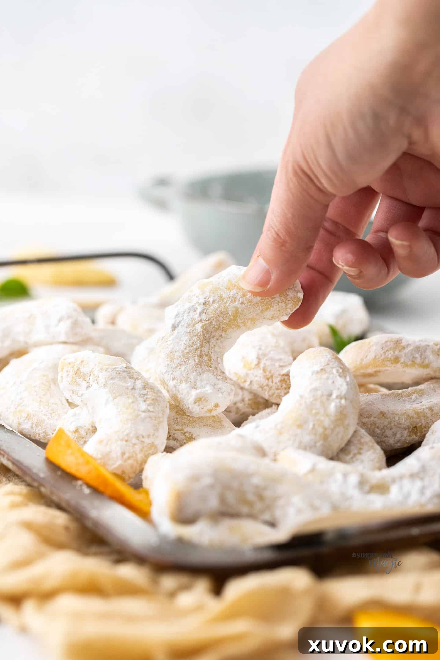 A hand reaching to pick up a golden orange almond crescent cookie from a metal baking tray, generously dusted with powdered sugar, highlighting its perfect shape and texture.