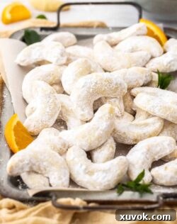 A batch of orange almond crescent cookies on a metal tray, dusted with powdered sugar.