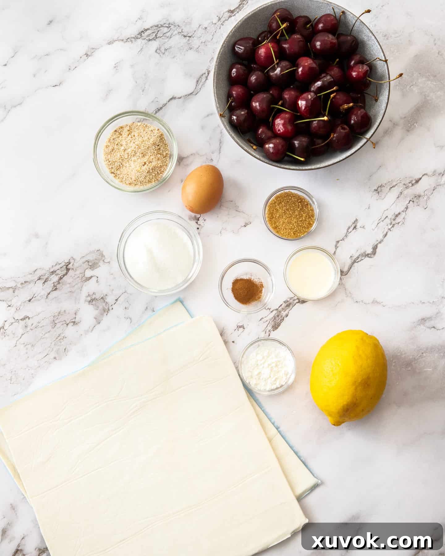 Ingredients for cherry strudel laid out on a marble surface, including fresh cherries, lemon, sugar, puff pastry, and spices.