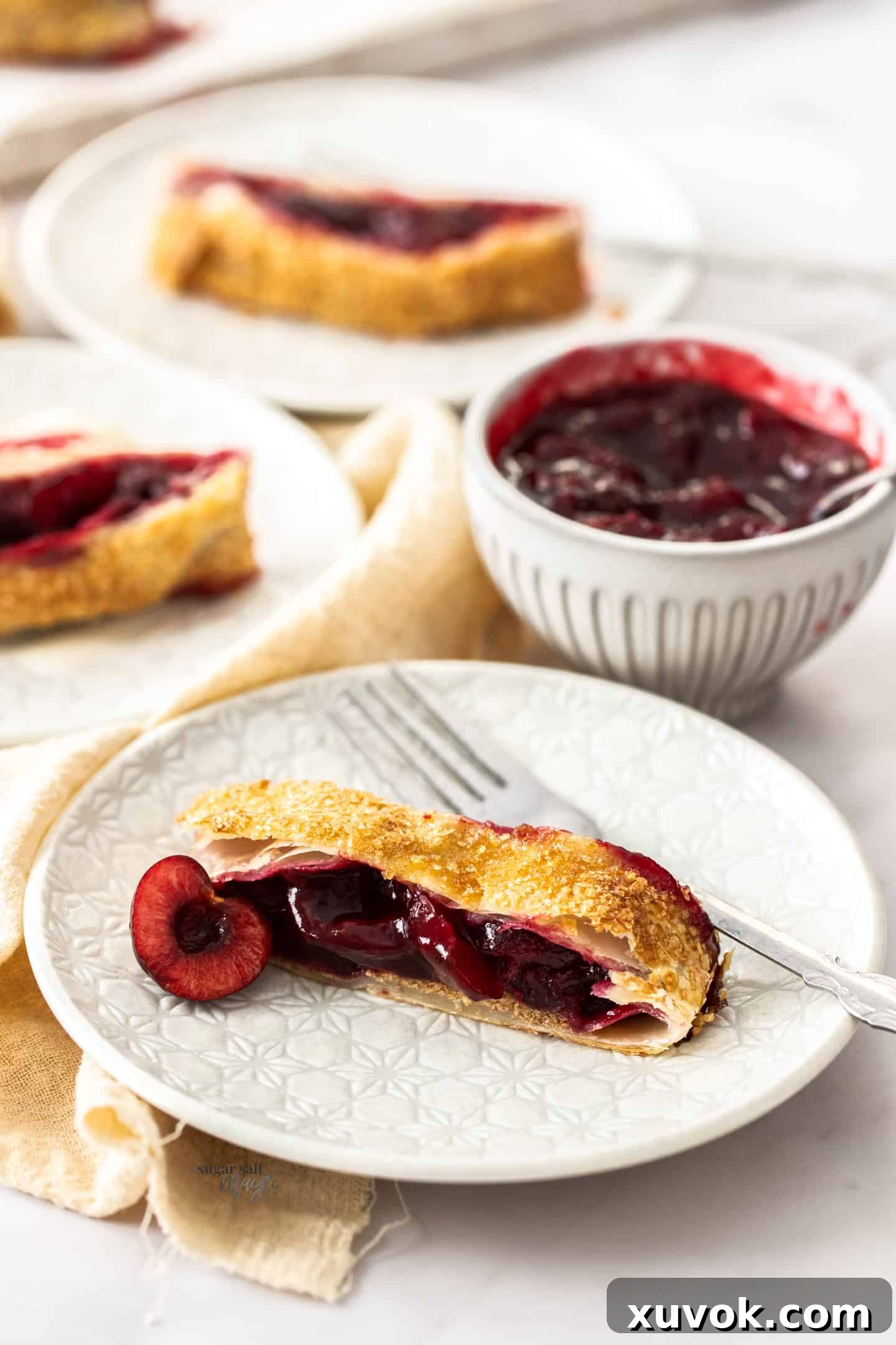 A slice of cherry strudel on a small white plate, showing the golden, flaky crust and red cherry filling.