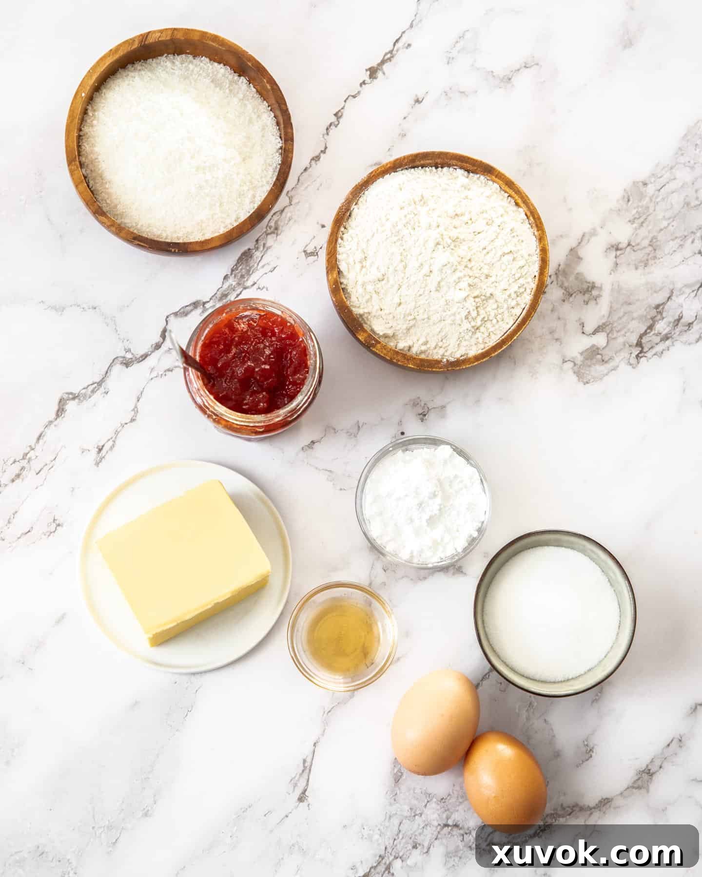 Ingredients for coconut tartlets laid out on a marble surface, including flour, butter, eggs, sugar, jam, vanilla, and desiccated coconut.