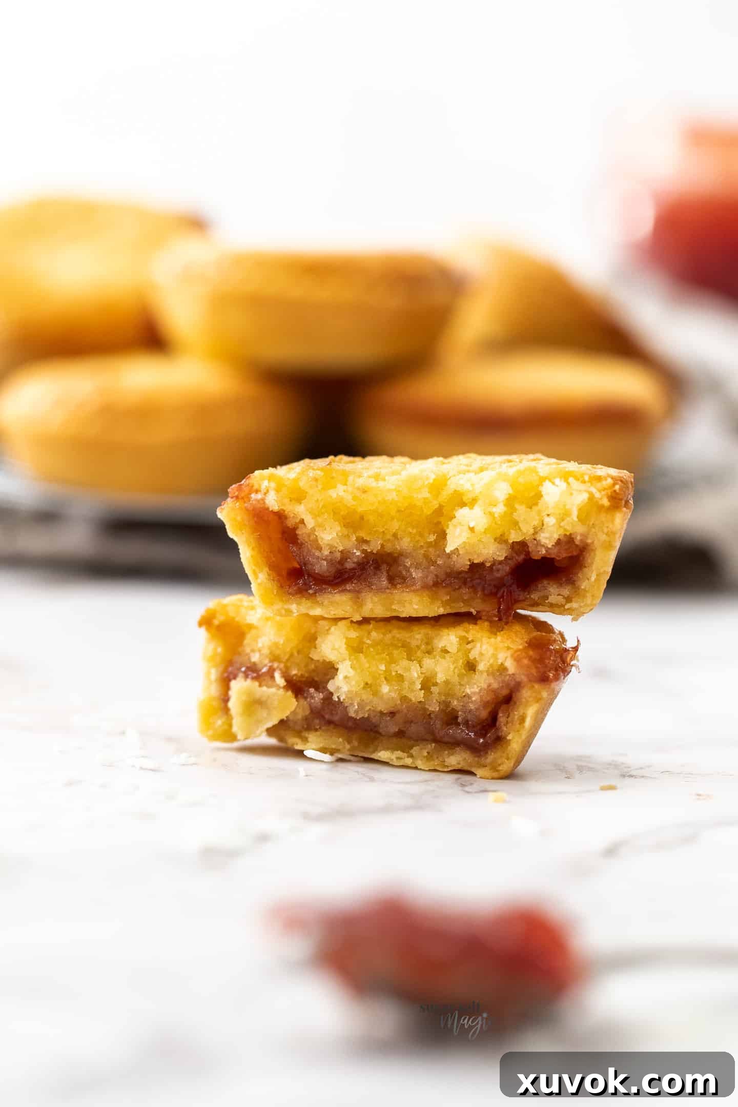 A coconut tartlet broken in half to reveal its jam and coconut layers, stacked on top of another whole tartlet.