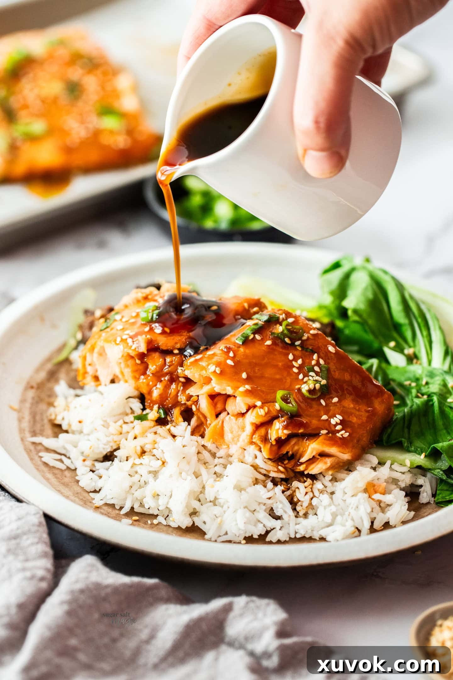 A jug of homemade teriyaki sauce elegantly drizzling over a perfectly oven-baked teriyaki glazed salmon fillet, served atop a bed of steamed rice.