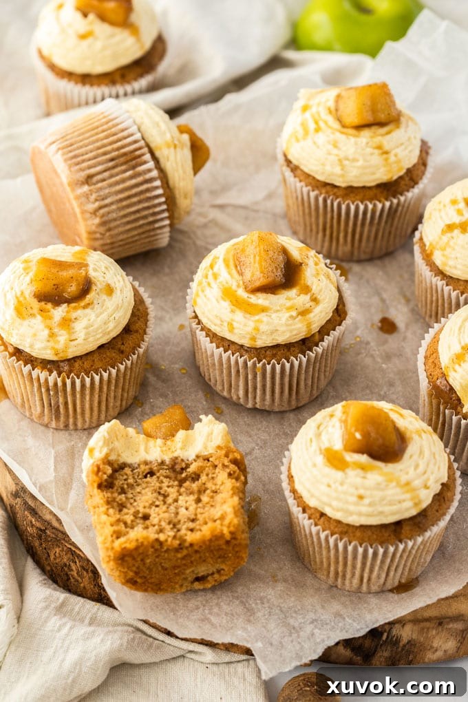 Apple pie cupcakes sitting on a wooden cake board, one is half eaten, showcasing fluffy cake and creamy frosting.
