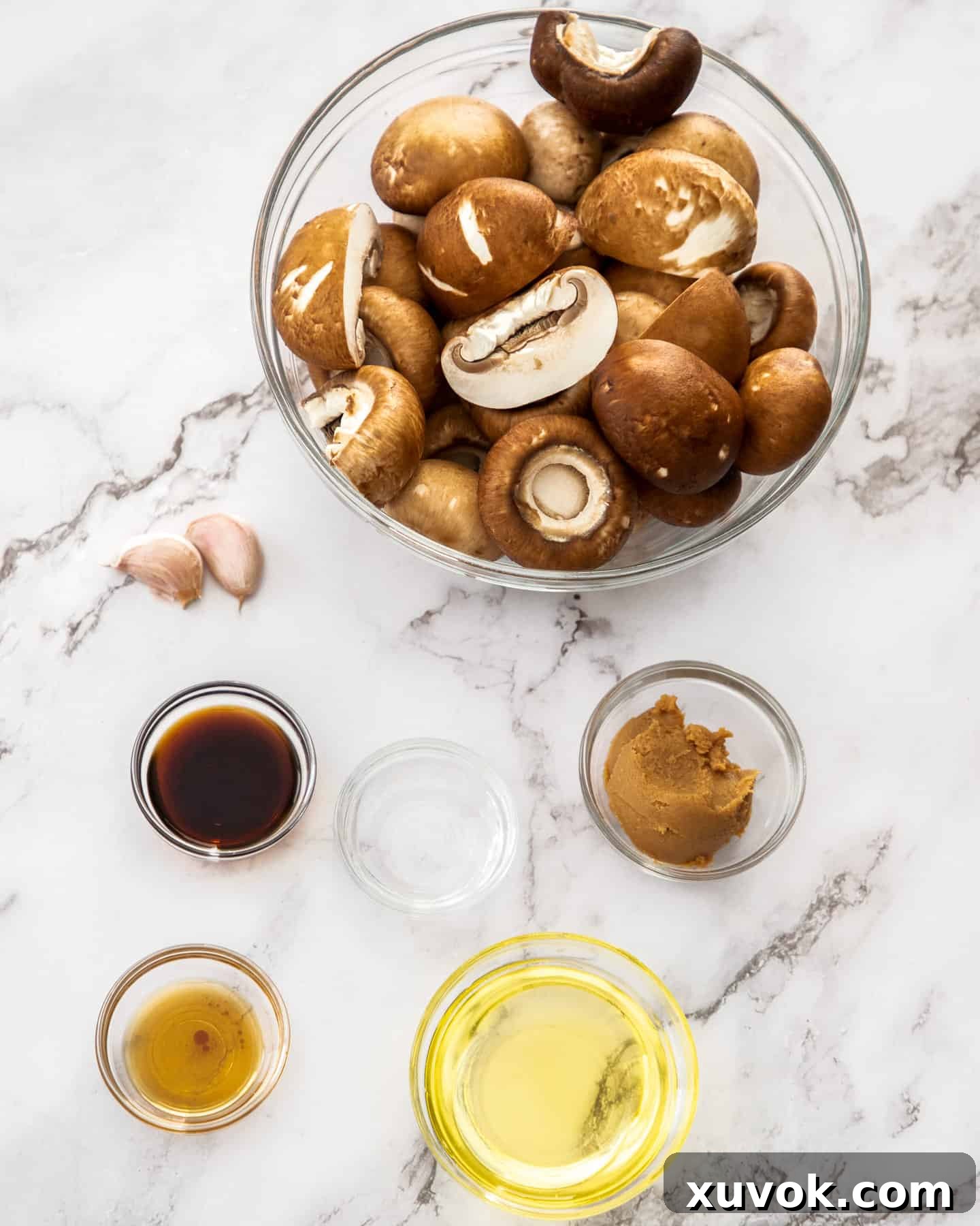 A collection of fresh ingredients for miso mushrooms, including whole mushrooms, miso paste, and garlic, arranged on a marble countertop.