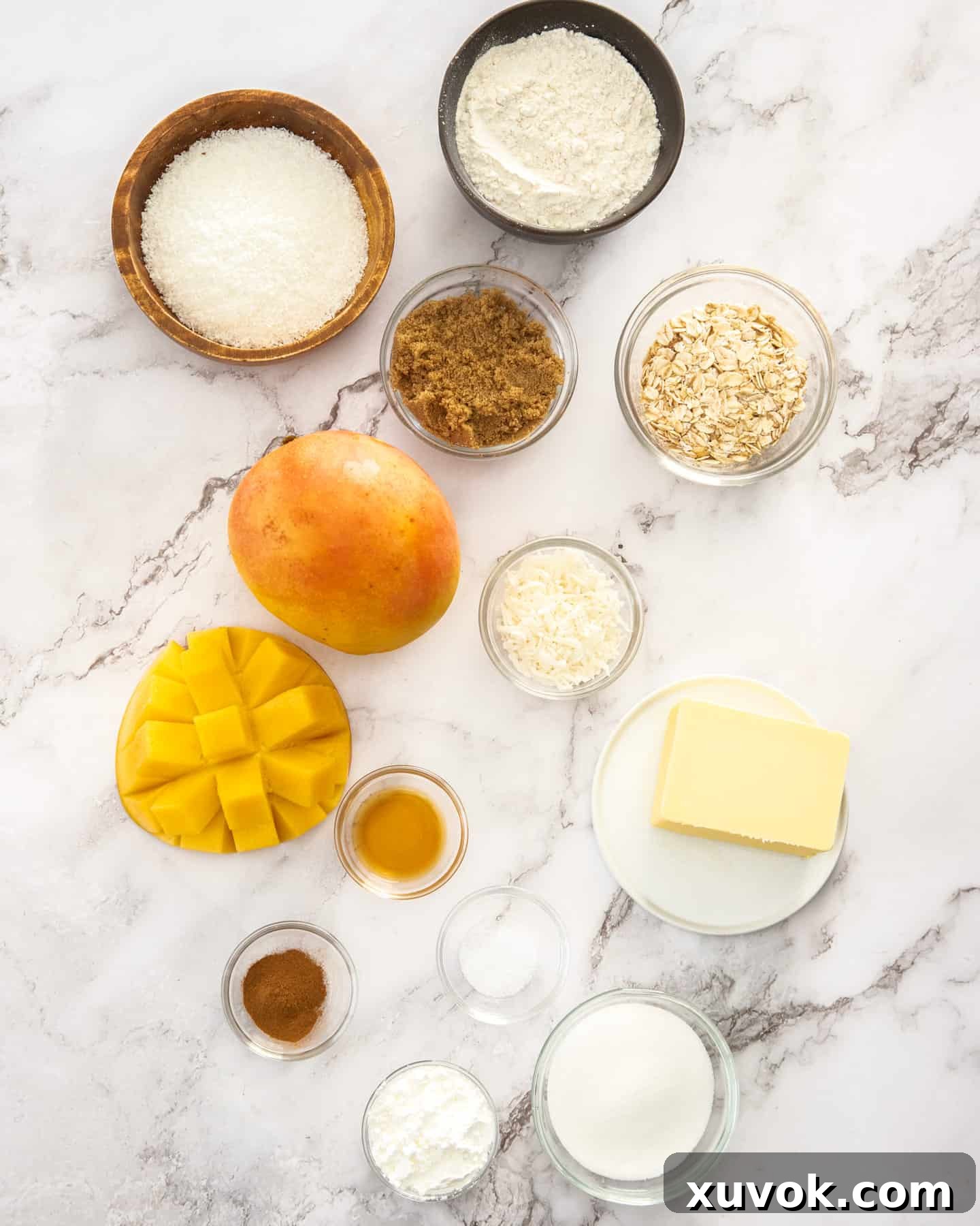 A thoughtfully arranged collection of fresh mangos, flour, oats, sugars, butter, coconut, vanilla extract, cinnamon, and cornstarch, neatly laid out on a pristine marble countertop, ready for baking this tropical crumble.