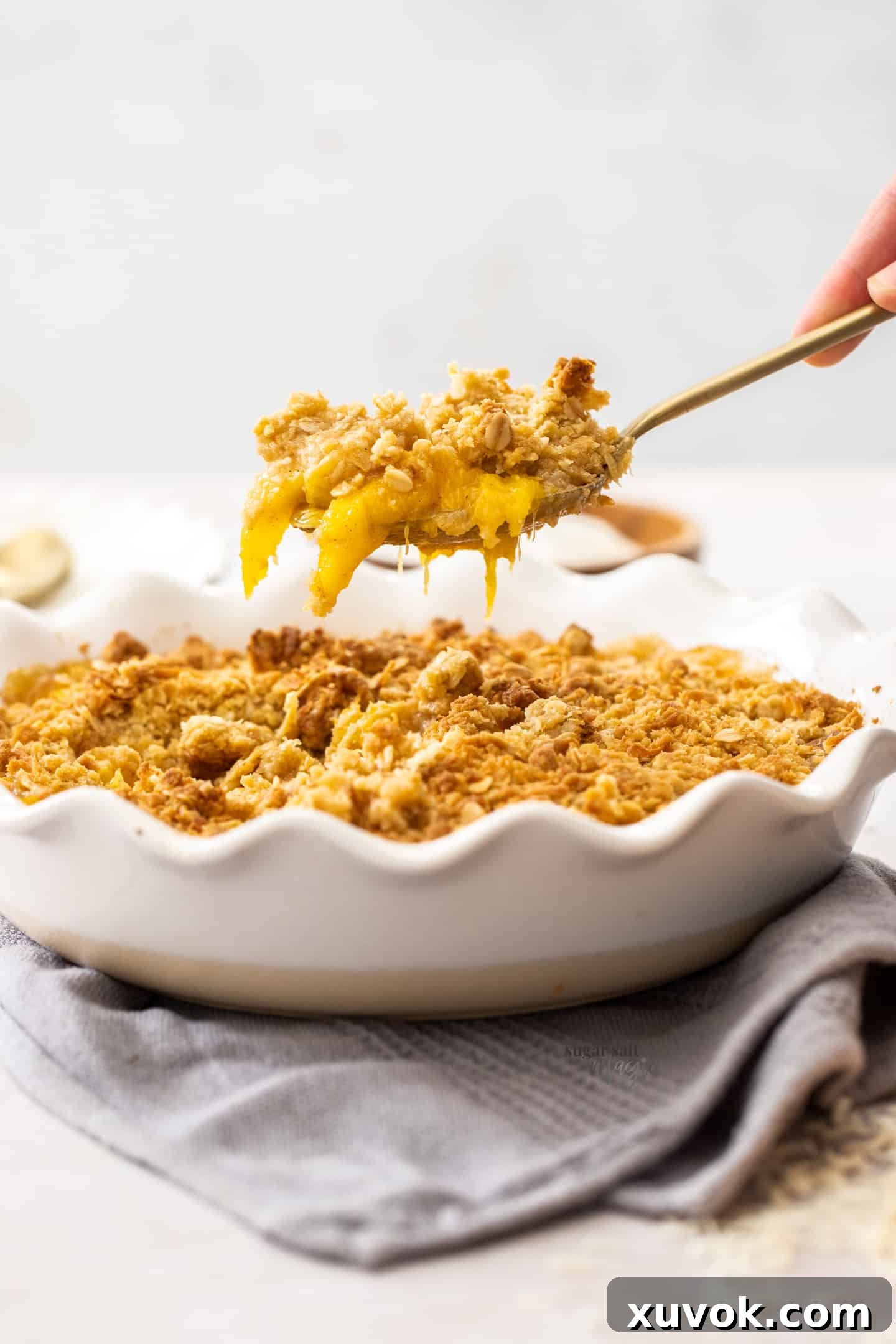 A golden spoon poised to scoop a portion of the freshly baked coconut mango crumble directly from the warm pie dish, highlighting its bubbling fruit filling and crisp, golden-brown topping.