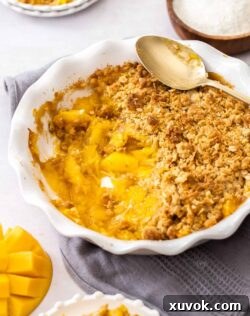A pie dish filled with golden-brown mango crumble, with a portion scooped out, ready to be served, capturing its inviting texture.