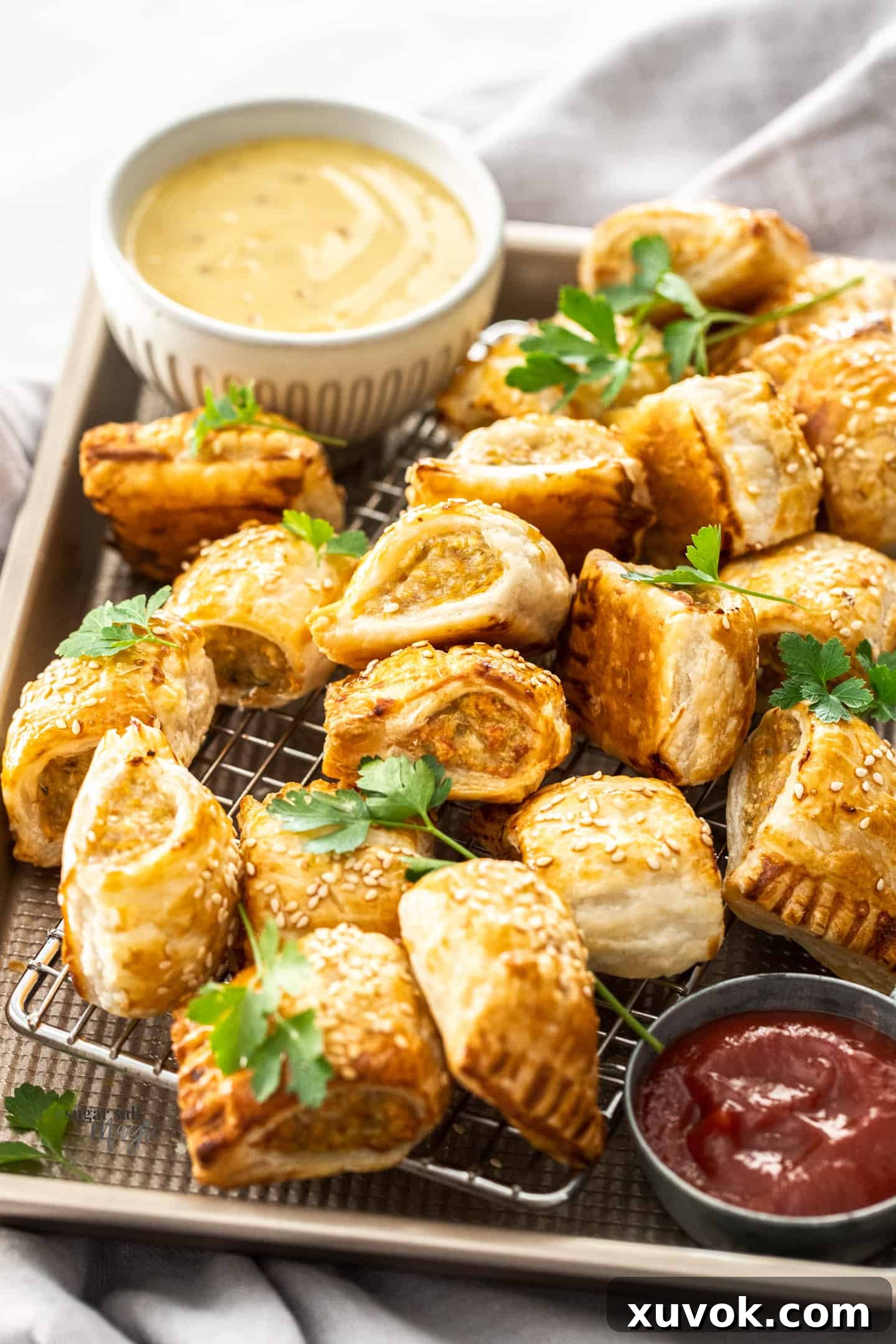 A close-up view of freshly baked chicken sausage rolls arranged in a baking tray with a bowl of dipping sauce blurred in the background.