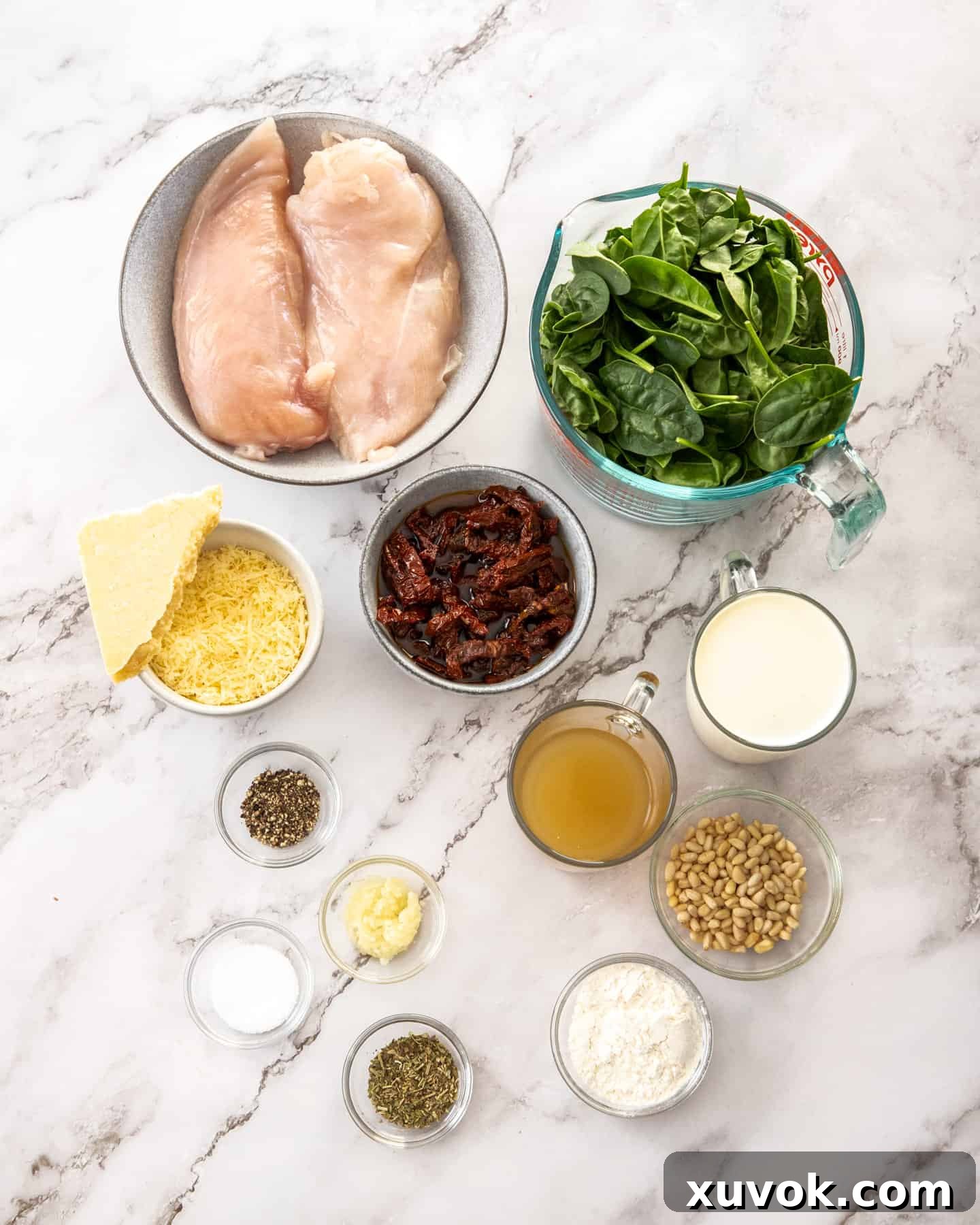 Ingredients for creamy Tuscan chicken laid out on a marble surface, including chicken breasts, spinach, sun-dried tomatoes, garlic, cream, and cheese.