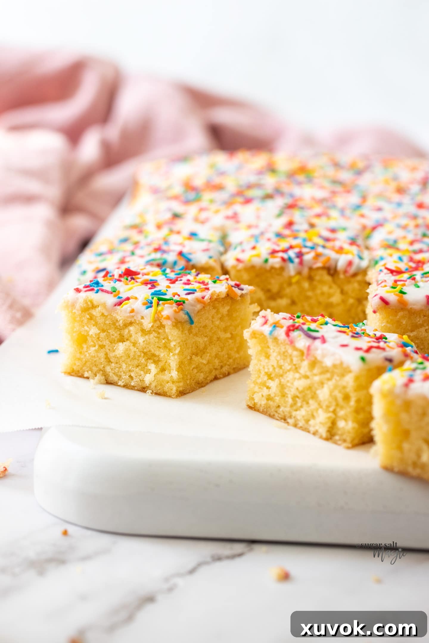 Squares of soft and fluffy vanilla tray bake cake topped with simple white icing and colorful sprinkles, ready for serving.