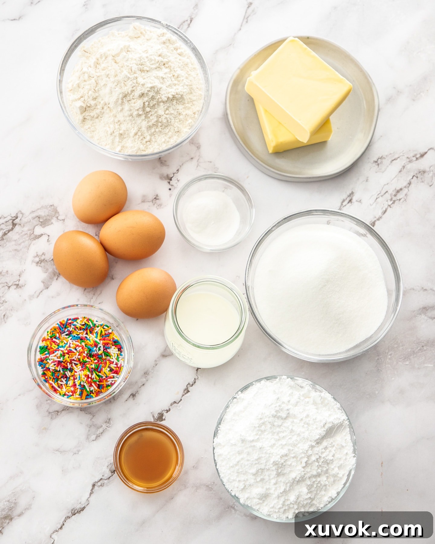 A beautifully arranged selection of fresh ingredients for baking vanilla tray bake, including flour, sugar, butter, eggs, milk, and vanilla extract, all resting on a elegant marble surface.