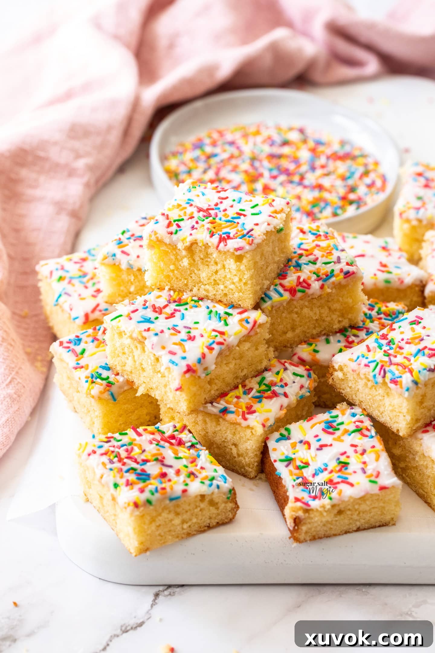 A beautifully composed stack of squares of vanilla sponge tray bake, generously topped with simple white icing and a scattering of colorful sprinkles, ready for enjoyment.