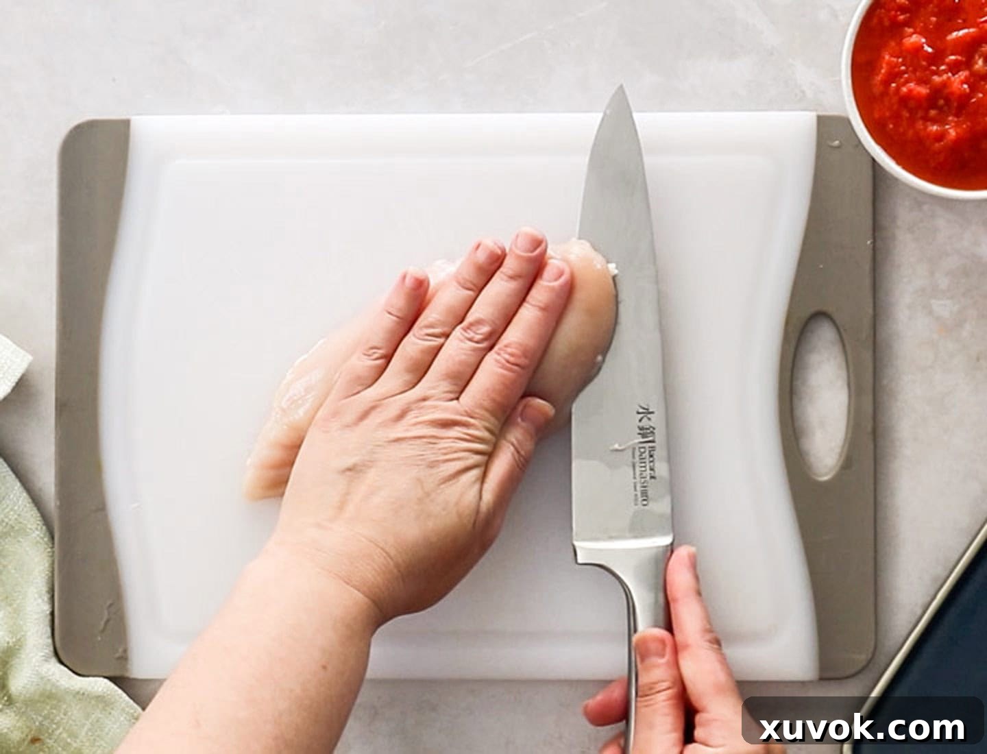 A hand holding down a chicken breast while a sharp knife slices it horizontally into thinner cutlets on a chopping board.