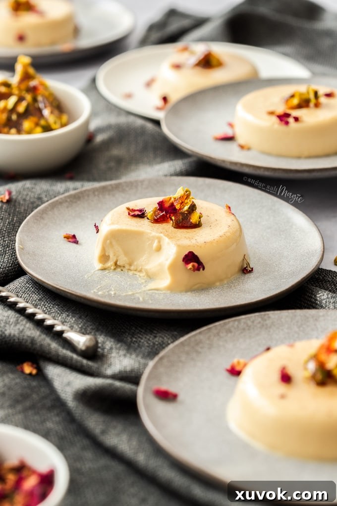 A single serving of Chai Panna Cotta on a grey plate, elegantly topped with crunchy pistachio toffee pieces and delicate rose petals, with a spoonful already taken revealing its creamy texture.
