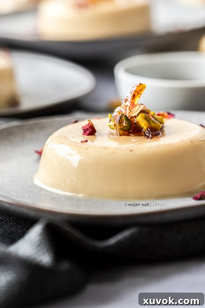 A detailed close-up of a single Chai Panna Cotta serving on a sophisticated grey plate, highlighting the glistening pistachio toffee shards and vibrant rose petals on its creamy surface.