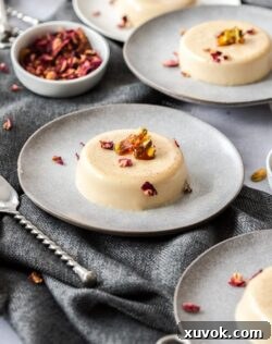 A creamy panna cotta dessert on a grey plate, topped with pistachio toffee and rose petals.