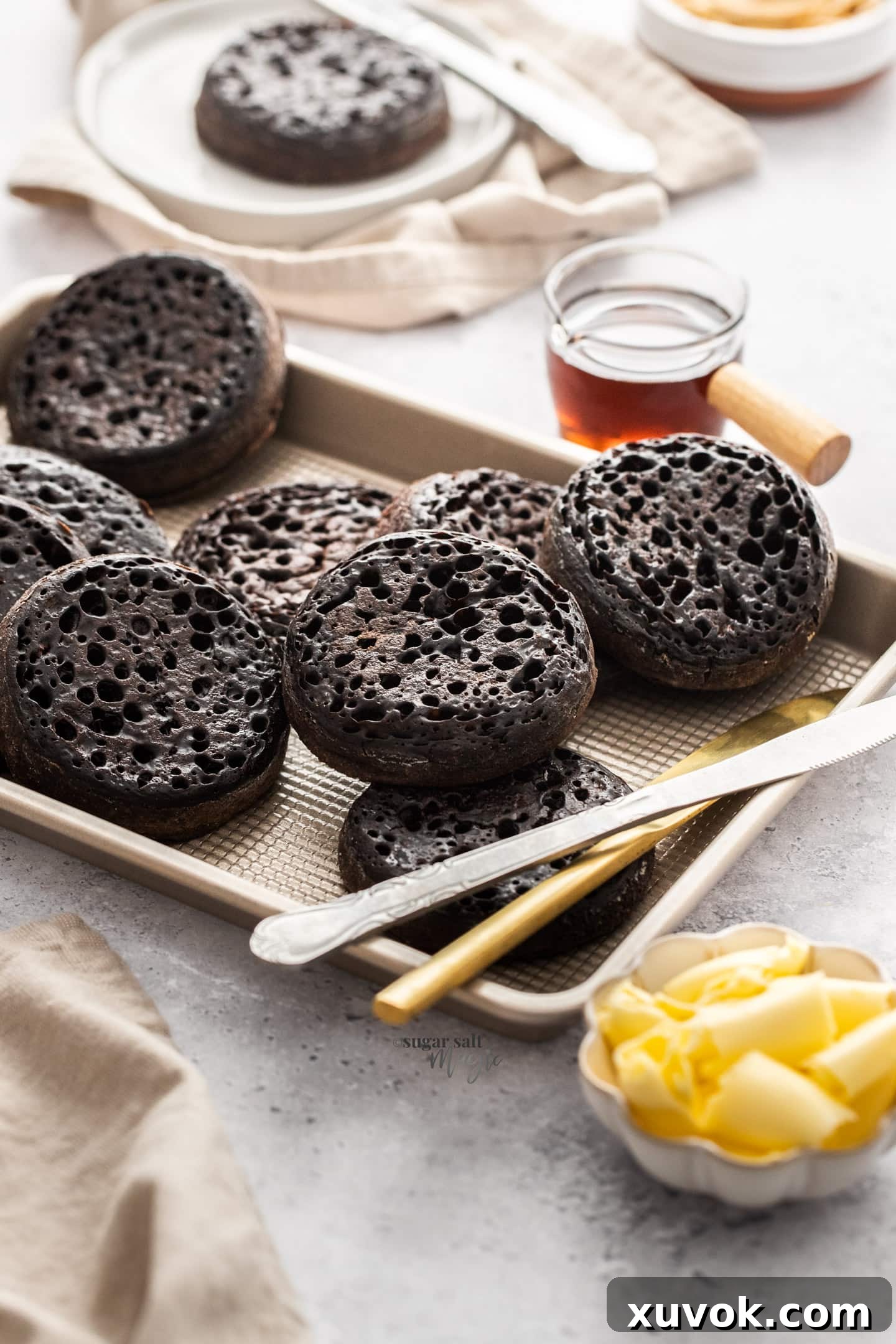 A batch of chocolate crumpets, next to a pot of maple syrup. Freshly made, golden brown with a rich chocolate hue, and characteristic holes on top, inviting toppings. The maple syrup adds a touch of sweetness.